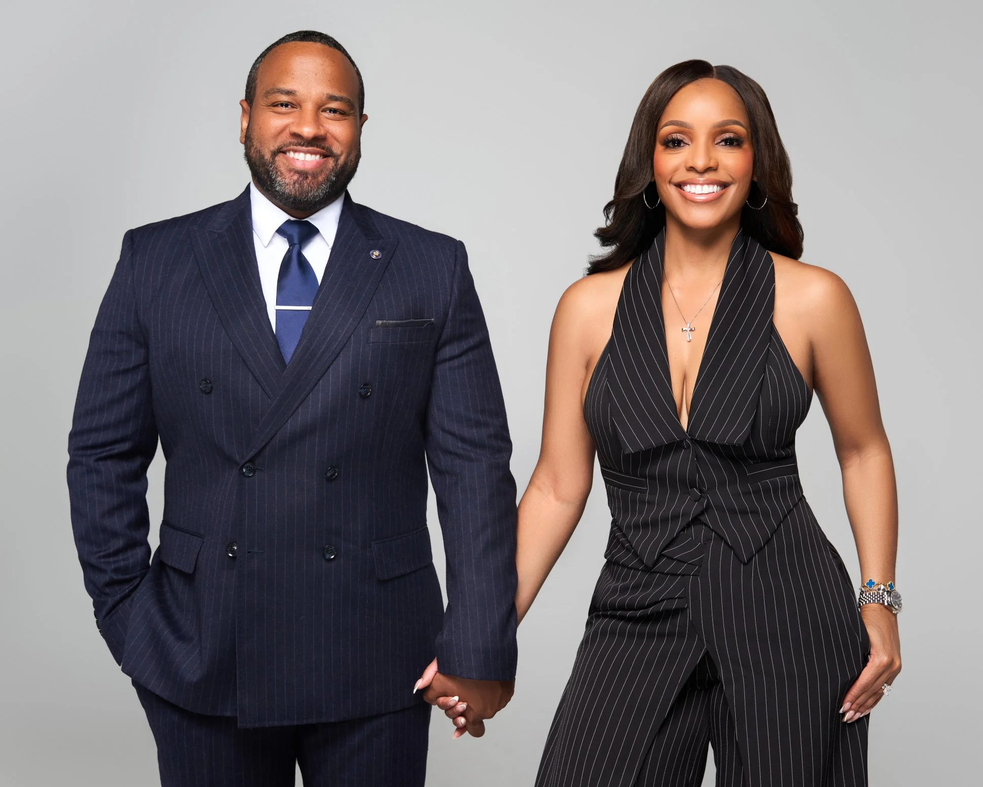 A man and woman holding hands, both dressed in formal clothing, standing against a plain gray background, smiling at the camera.
