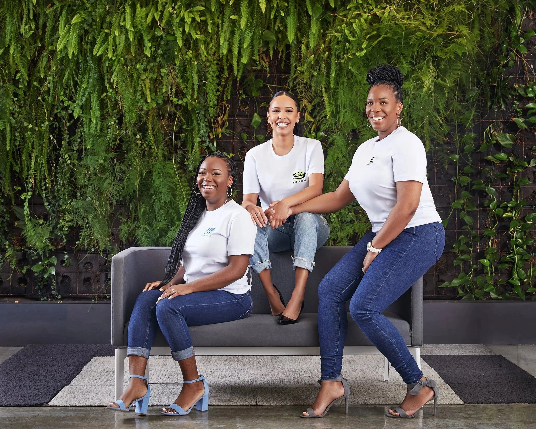 Three women wearing white shirts and jeans, smiling and sitting on and near a gray sofa in front of a green wall with plants.