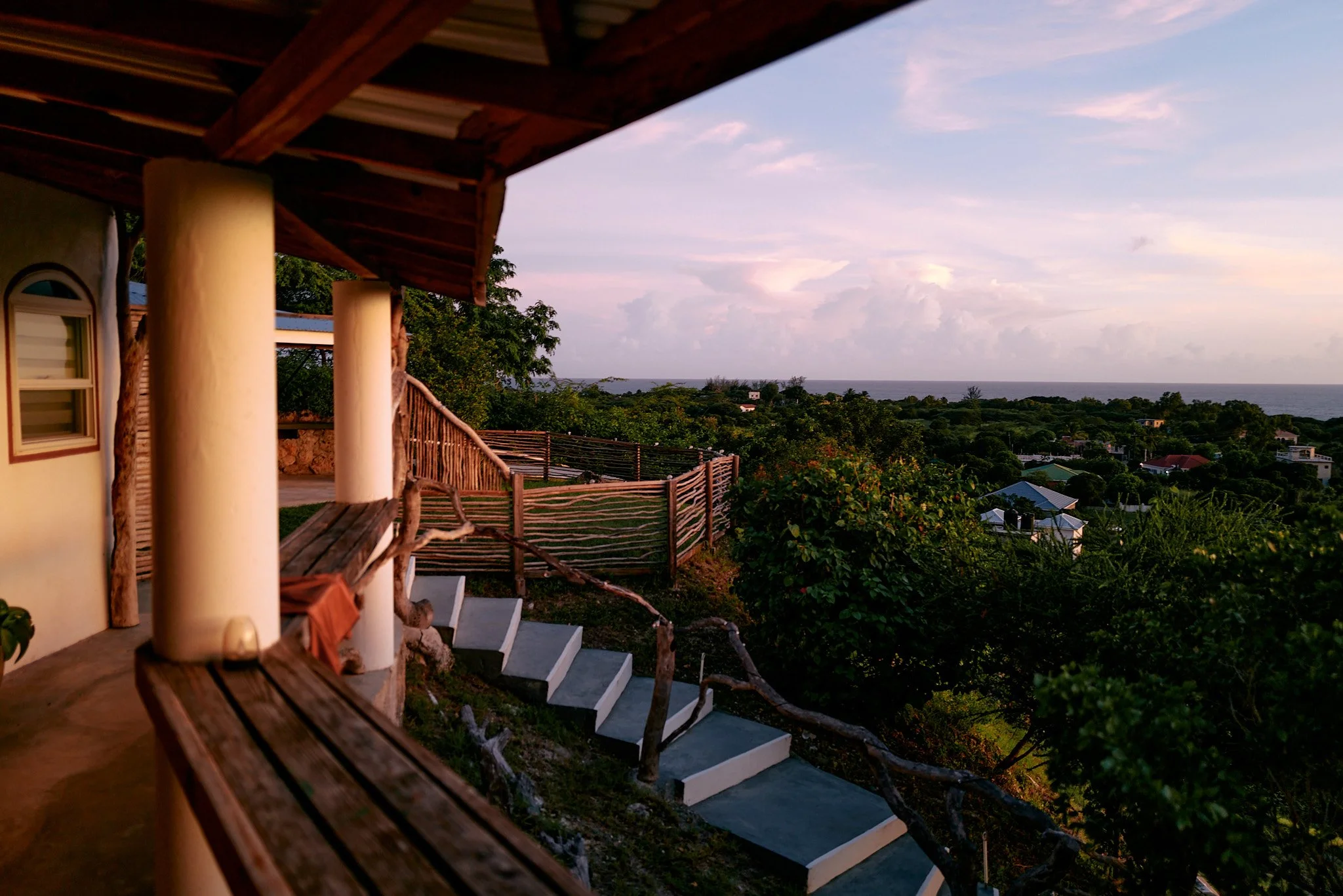 A view from a porch with a wooden railing overlooking a lush green landscape towards the ocean with cloudy sky at sunset in Treasure beach, Jamaica .