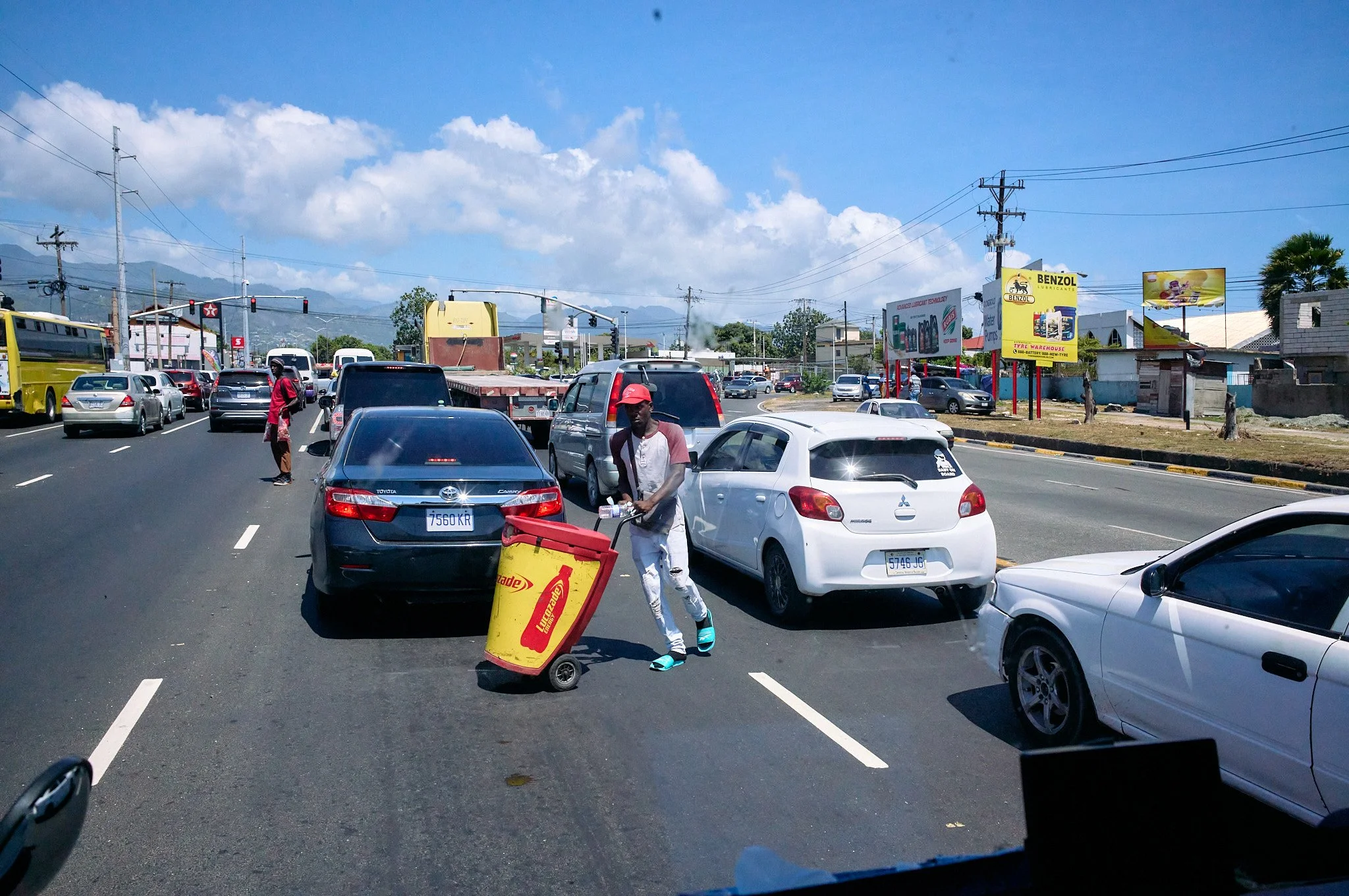 Traffic jam on a busy road with cars and buses moving slowly under a partly cloudy sky; a man wearing a red cap and a light shirt pushes a yellow trash bin in Kingston Jamaica