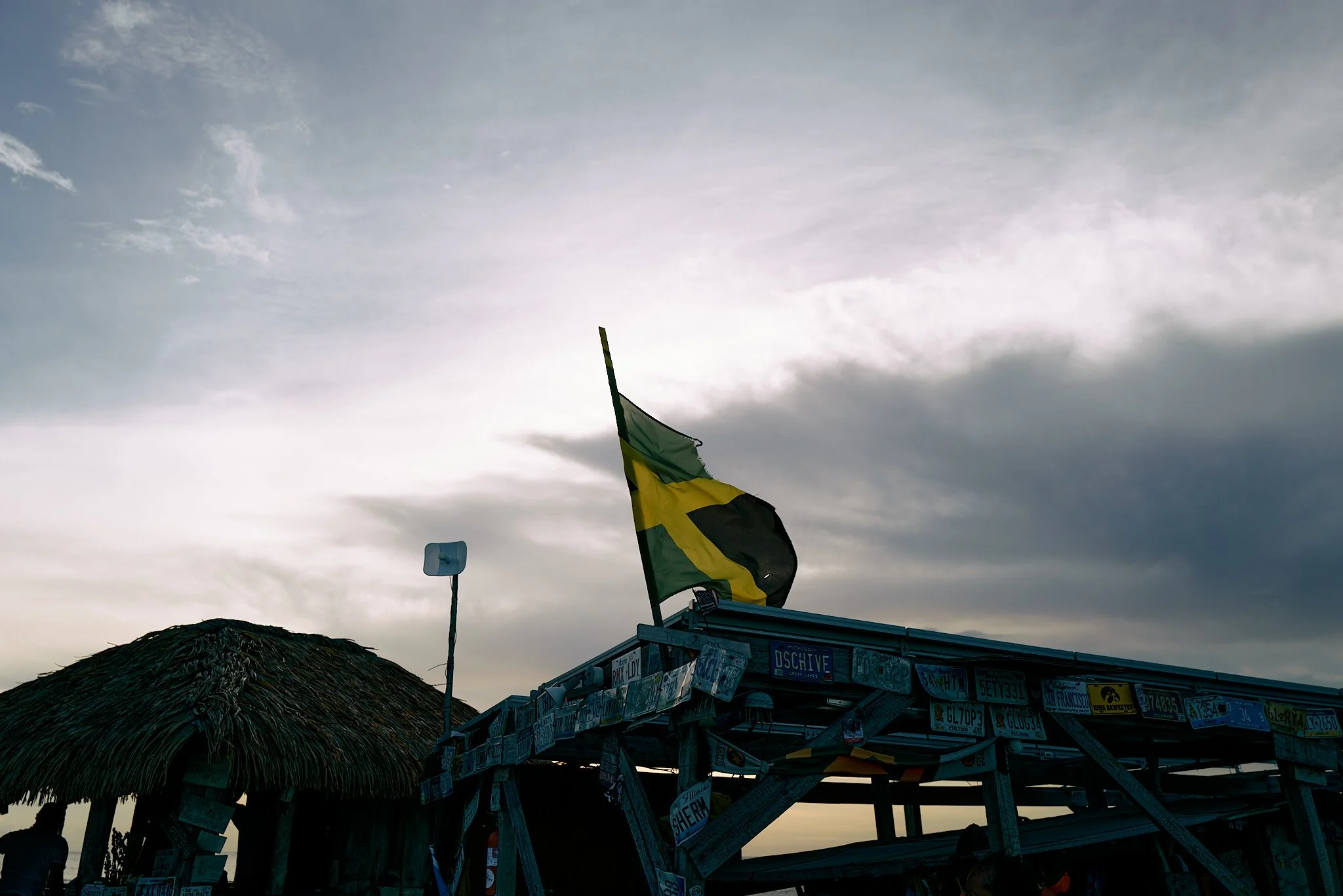 A Jamaican flag with green, yellow, and black colors flying over a structure decorated with license plates, against a cloudy sky at sunset or dusk.