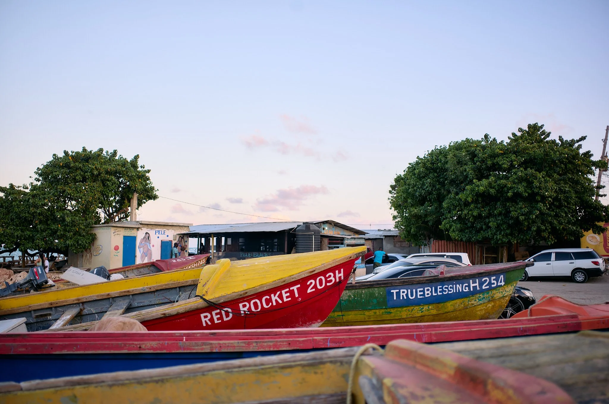 Colorful boats parked on shore, with license plates 'RED ROCKET 203H' and 'TRUE BLESSING H.254,' near trees and parked cars, small buildings, and a clear sky in Hellshire Beach, Jamaica