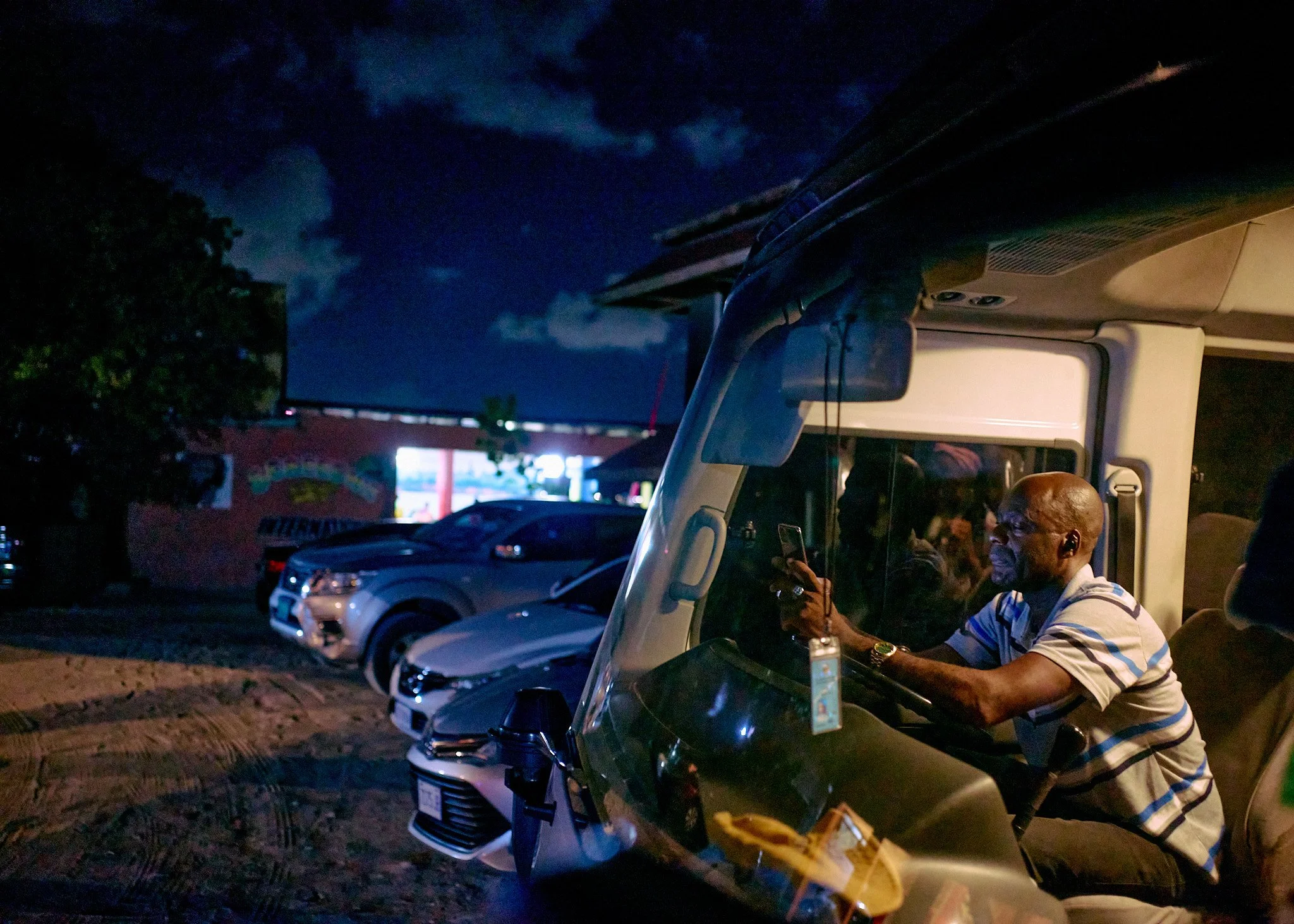A man driving a vehicle at night, looking at his phone, with parked cars and a building with bright lights in the background.