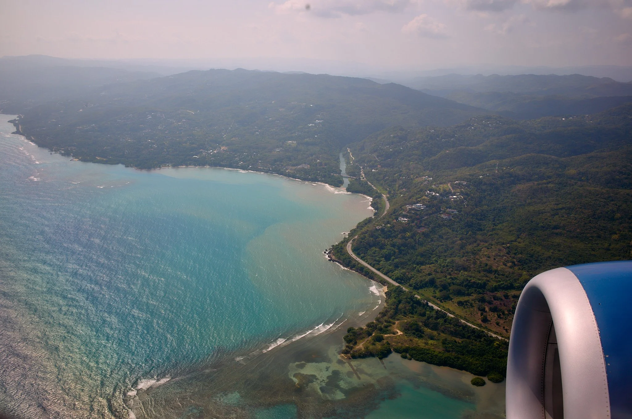 Aerial view from an airplane showing a large body of water with light blue and turquoise hues, surrounded by green hilly terrain with a winding road along the coast, and a partial view of the airplane engine in the bottom right corner.