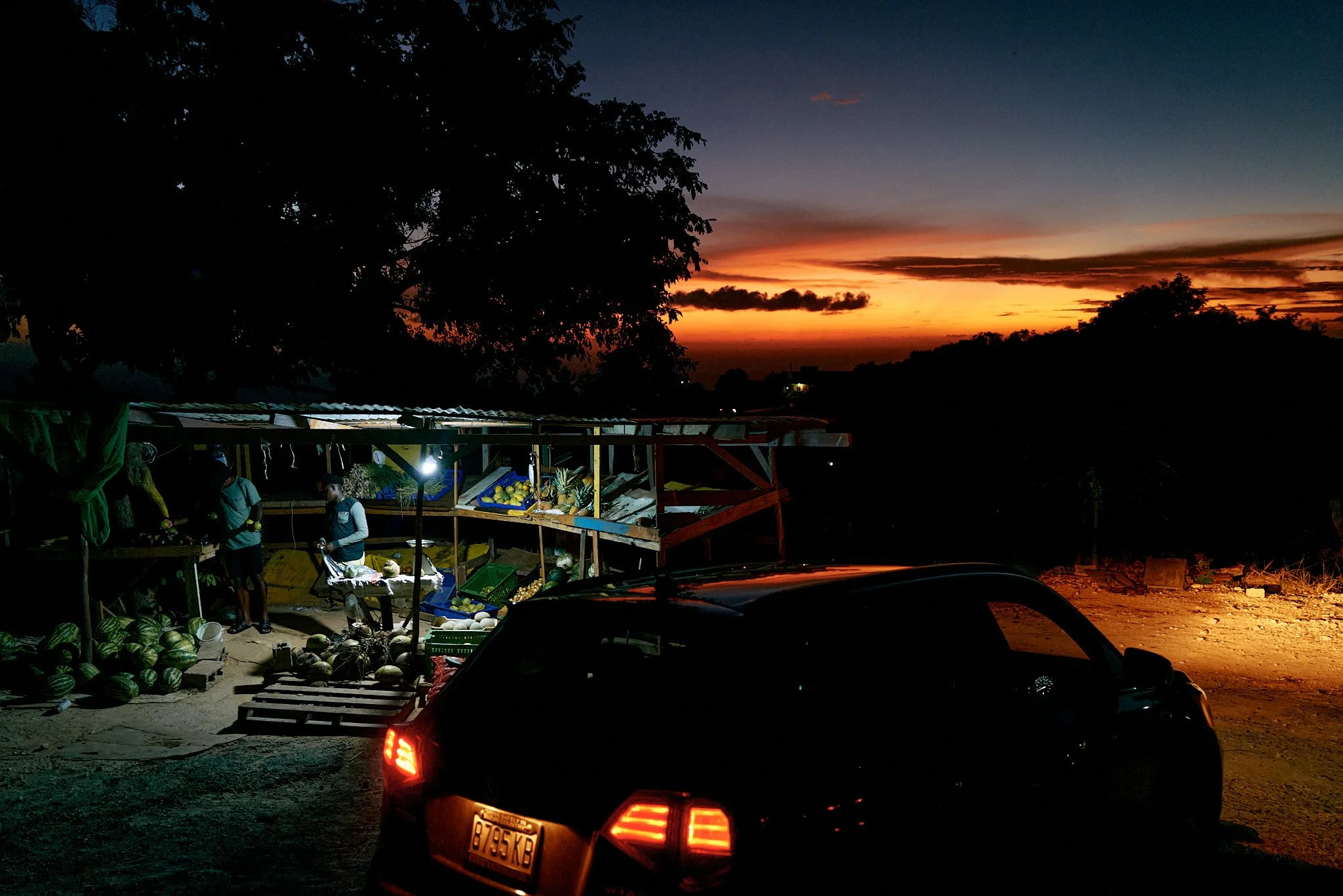 A roadside fruit stand at sunset with a dark car in the foreground, silhouetted trees, and a colorful sky with an orange and blue sunset.