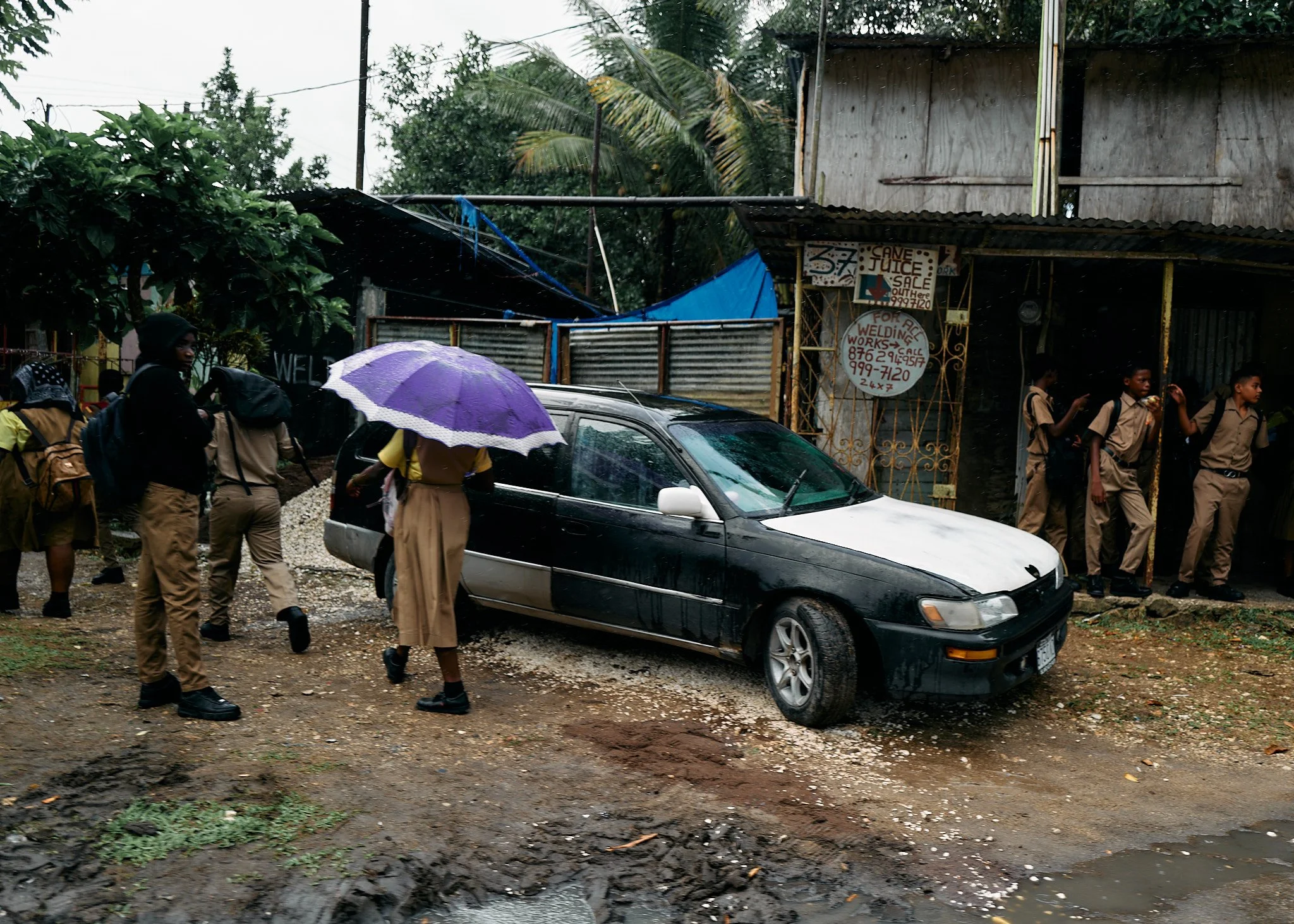 A group of students in school uniforms walking past a black and white car parked on a muddy road, with some students holding umbrellas in the rain, and a small storefront with signs in the background in Jamaica