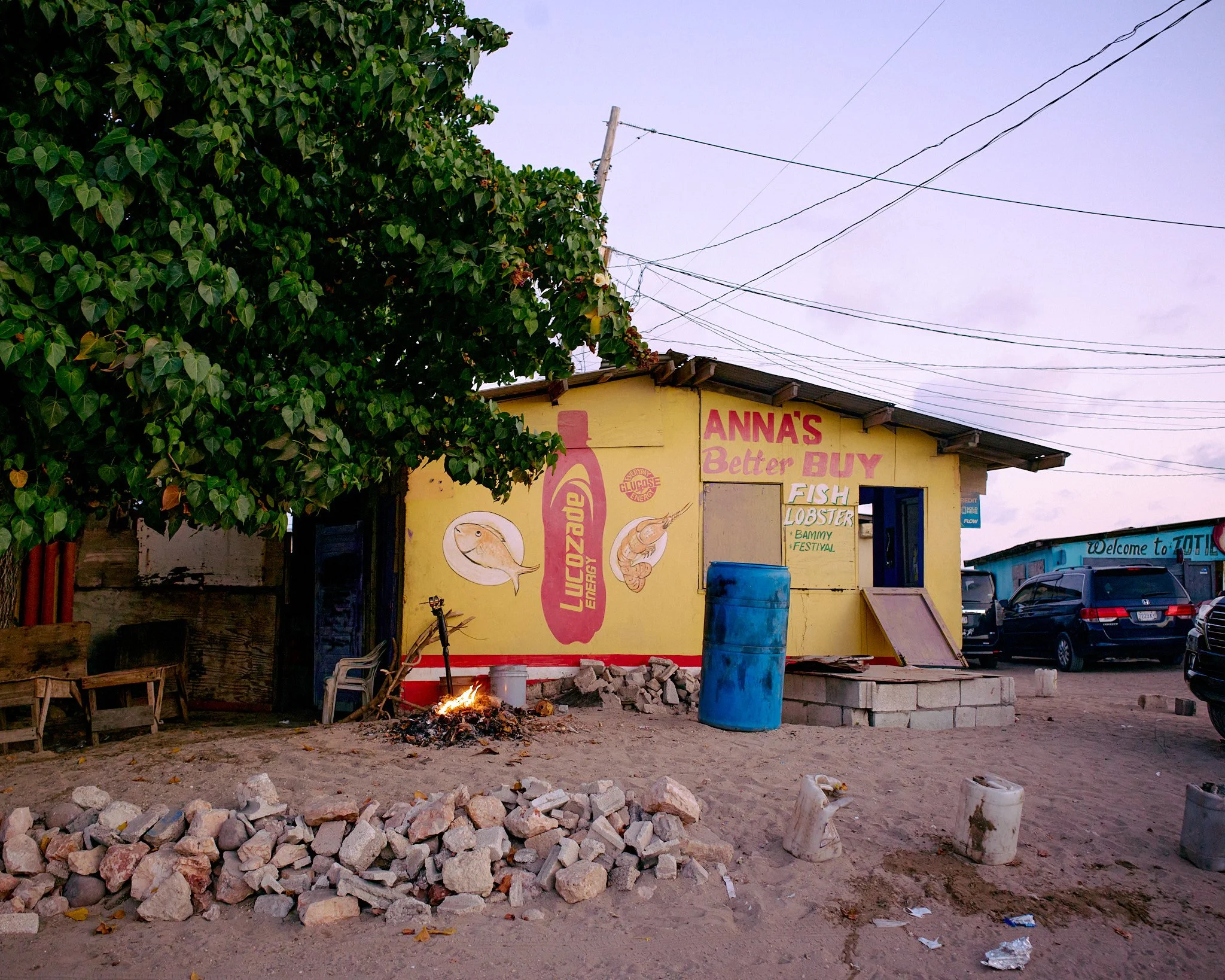 A small yellow building with signs for a seafood market, including fish and lobster, and a large bottle painted on the wall. The building has an open door, a blue barrel outside, and a small fire burning on the dirt ground in front. There are parked 