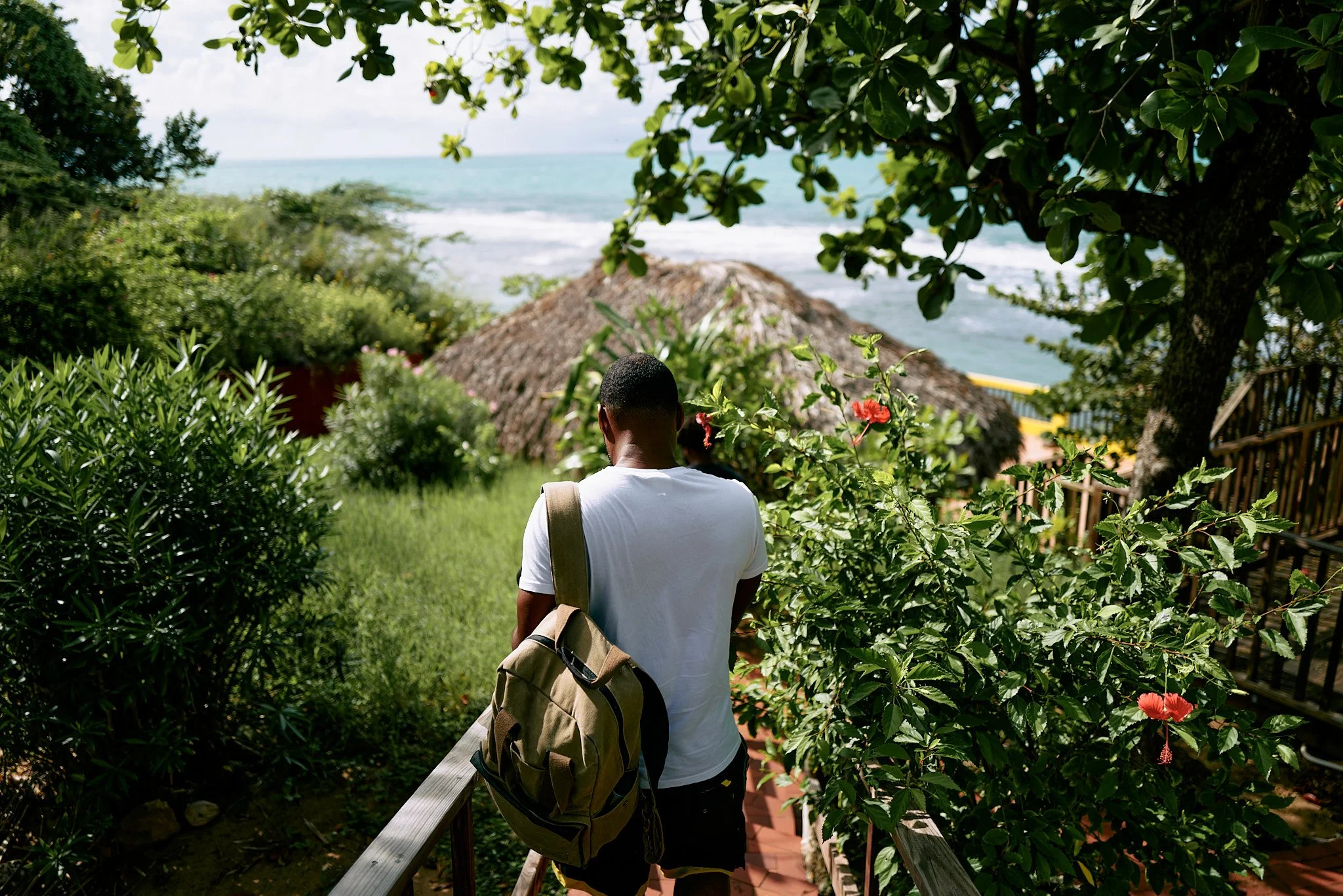 A man with a backpack walking down a wooden pathway surrounded by green bushes and trees. Jamaican waters in the background, there is a thatched roof hut near the ocean with waves and a cloudy sky.