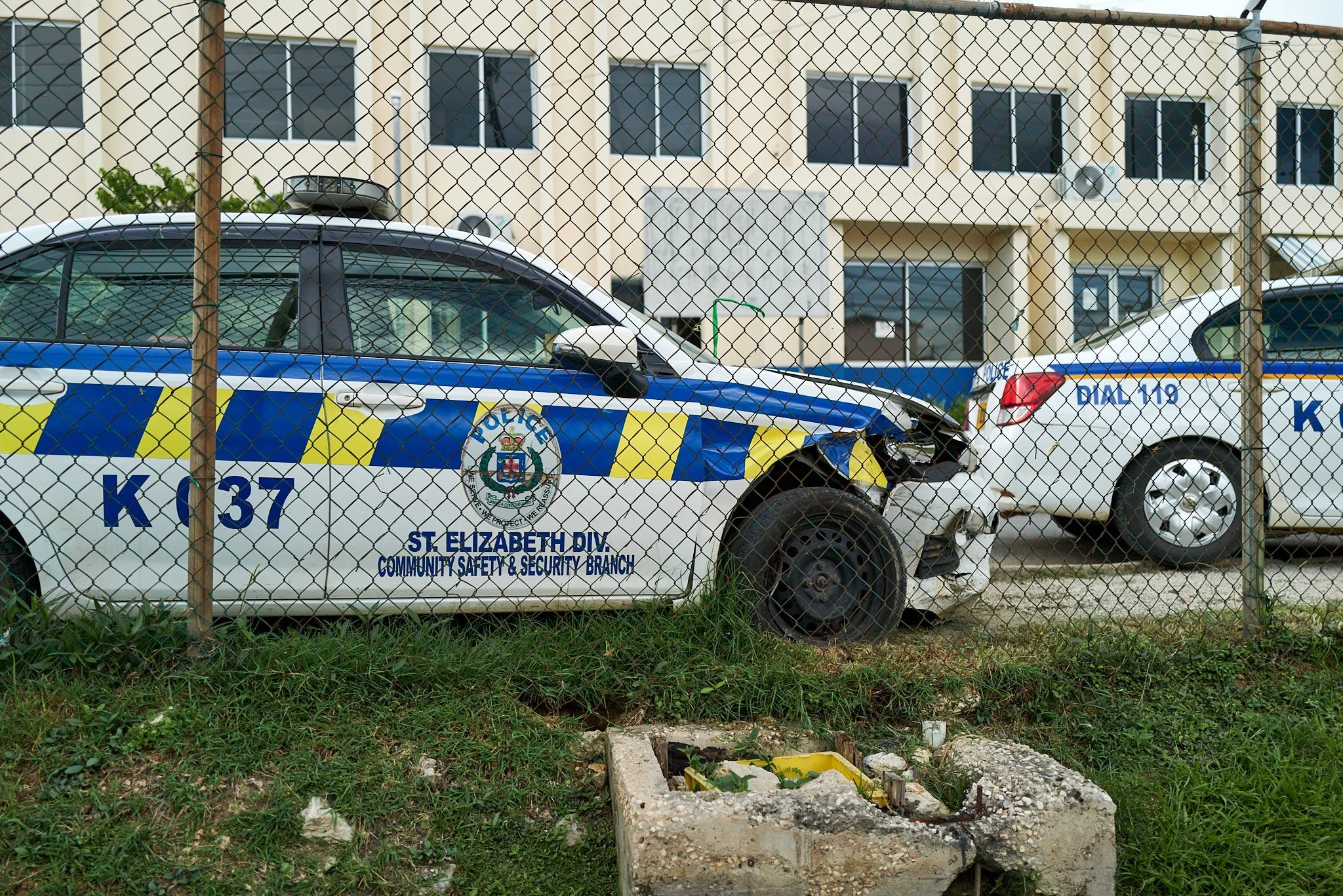 Damaged Jamaican police cars parked behind a chain-link fence, with a building in the background.