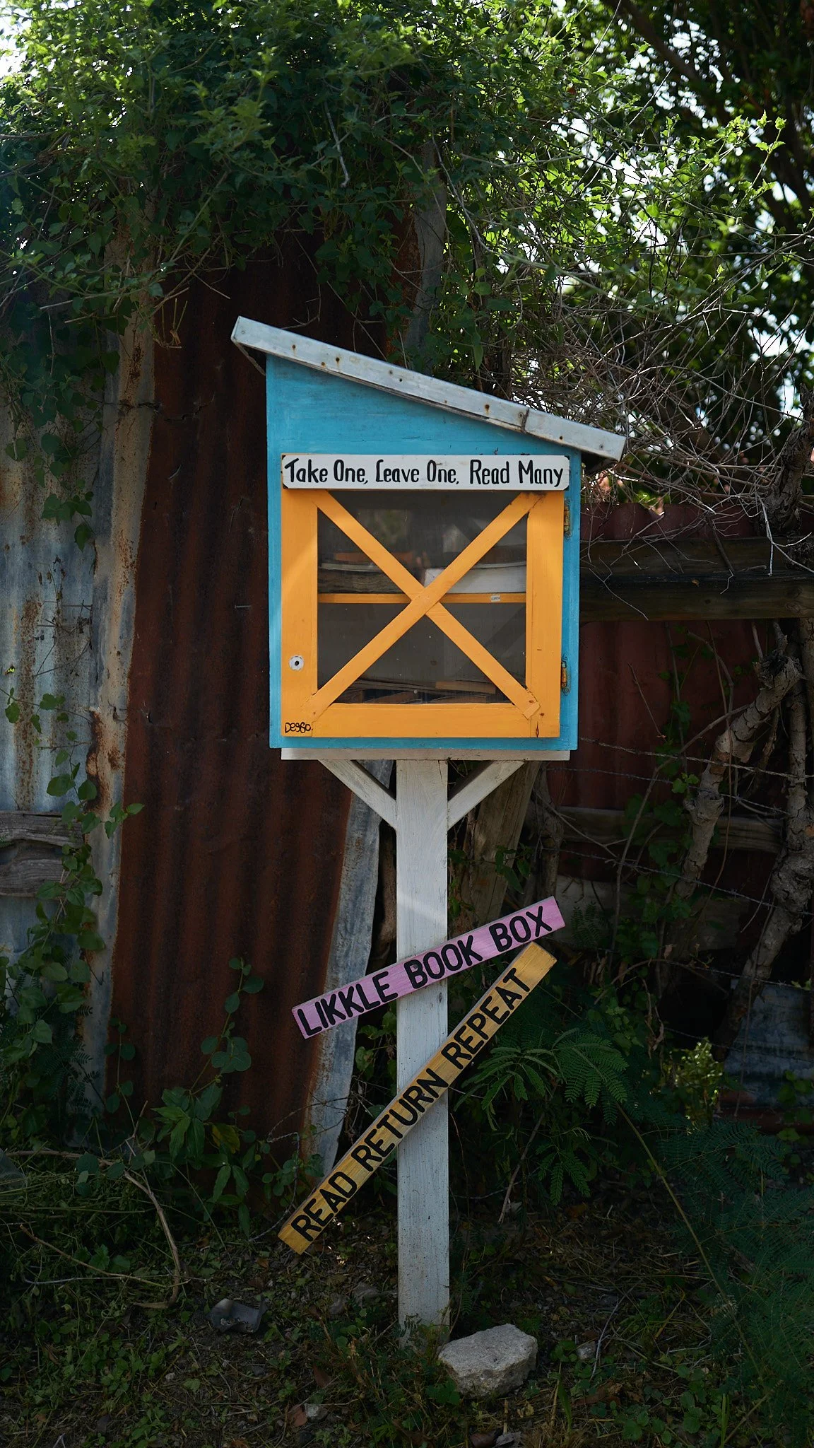 A decorative outdoor book exchange box with a colorful roof, a sign saying "Take One, Leave One, Read Many," and a smaller sign below that reads "Likkle Book Box."