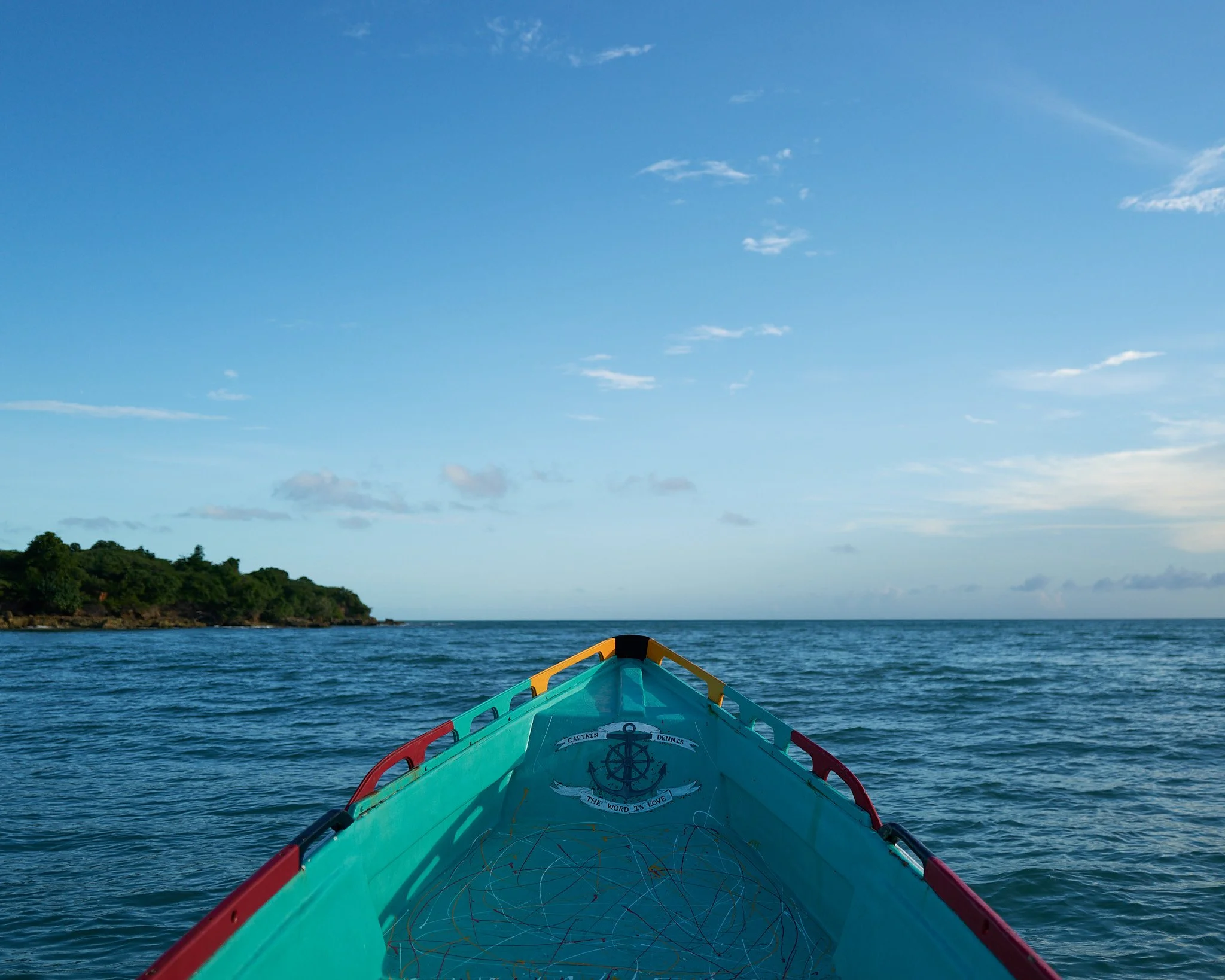 View from the front of a small boat on the ocean, with a distant green island on the left and a partly cloudy blue sky above in Treasure Beach Jamaica by Matt Ellis