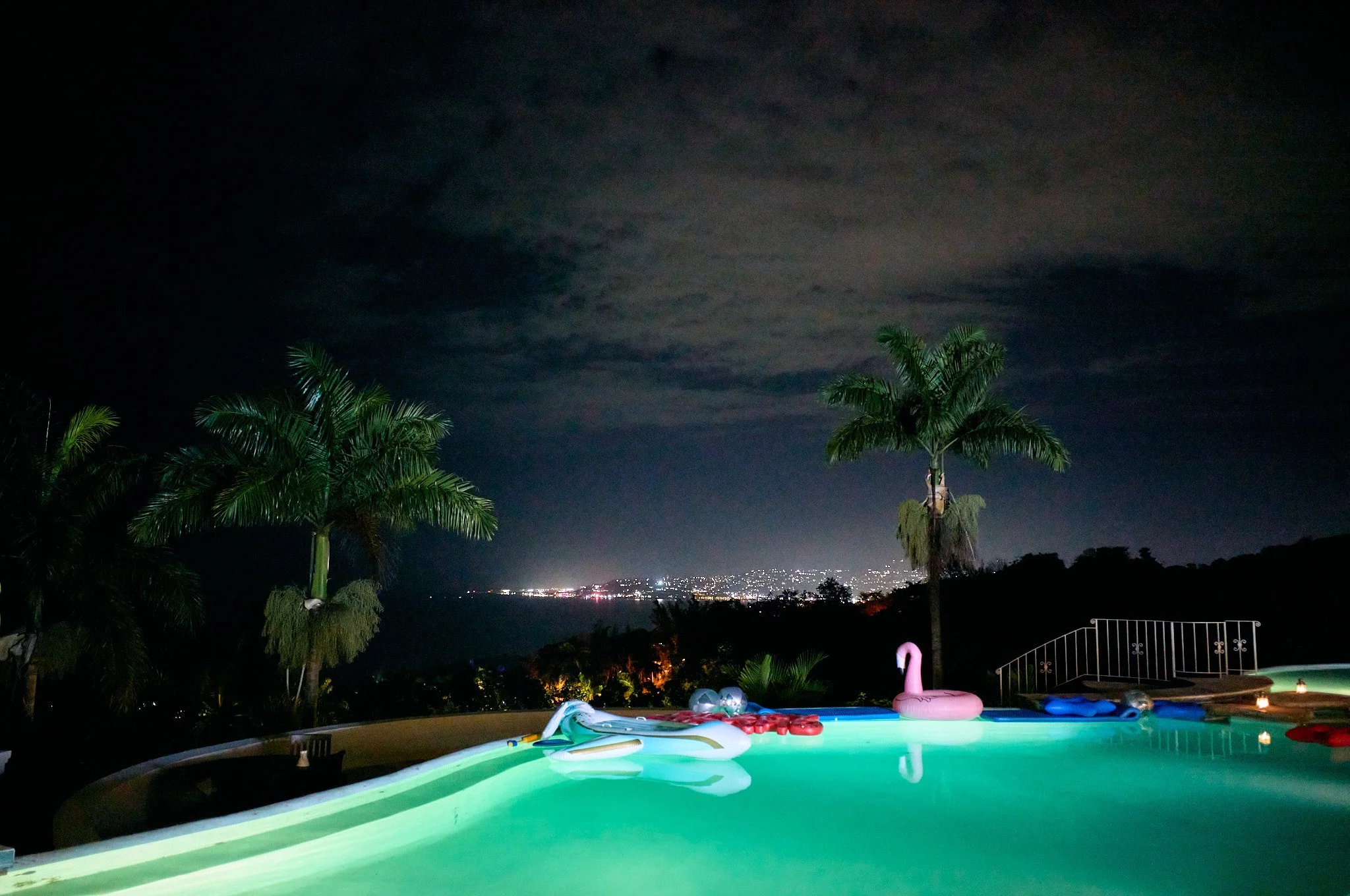 Nighttime view of a swimming pool with inflatable floats, including a pink flamingo and a white dolphin, surrounded by palm trees and overlooking the Montego Bay city skyline in the distance.