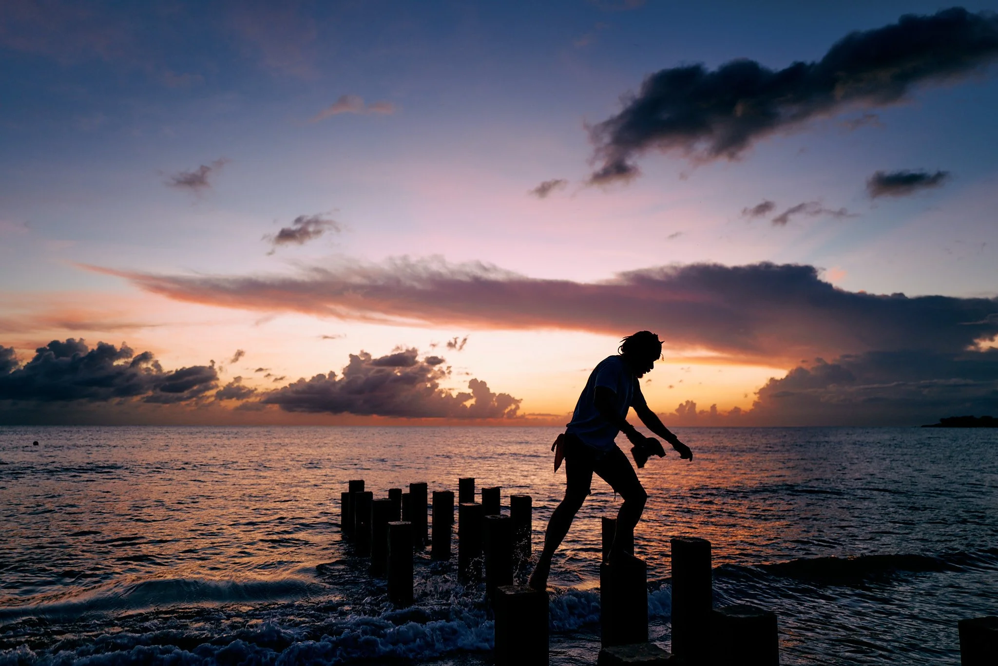Person walking on wooden posts in the ocean at sunset.