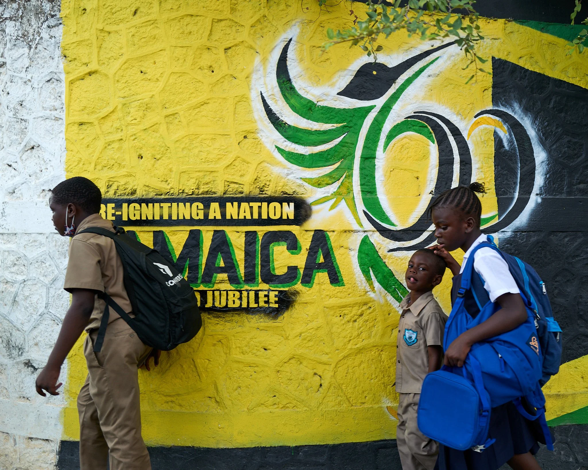 Three students in school uniforms walking past a bright yellow wall with a mural that includes the Jamaican flag and the words 'Pre-Igniting a Nation Jamaica Jubilee'.