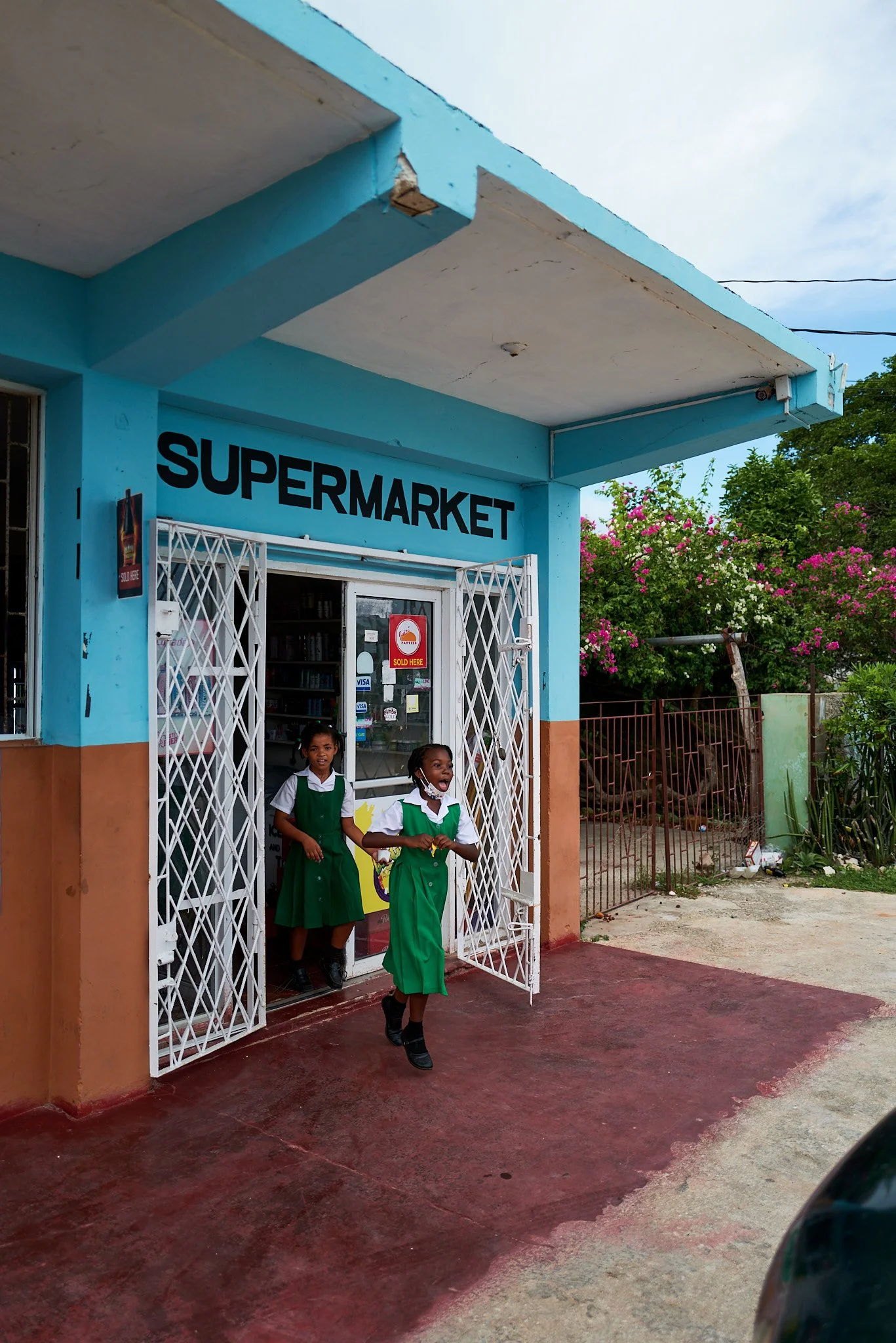 Two girls in green and white school uniforms exit a small blue supermarket with a red and brown front. The girl in front is smiling and holding hands with the girl behind her. There are pink flowering bushes to the right.