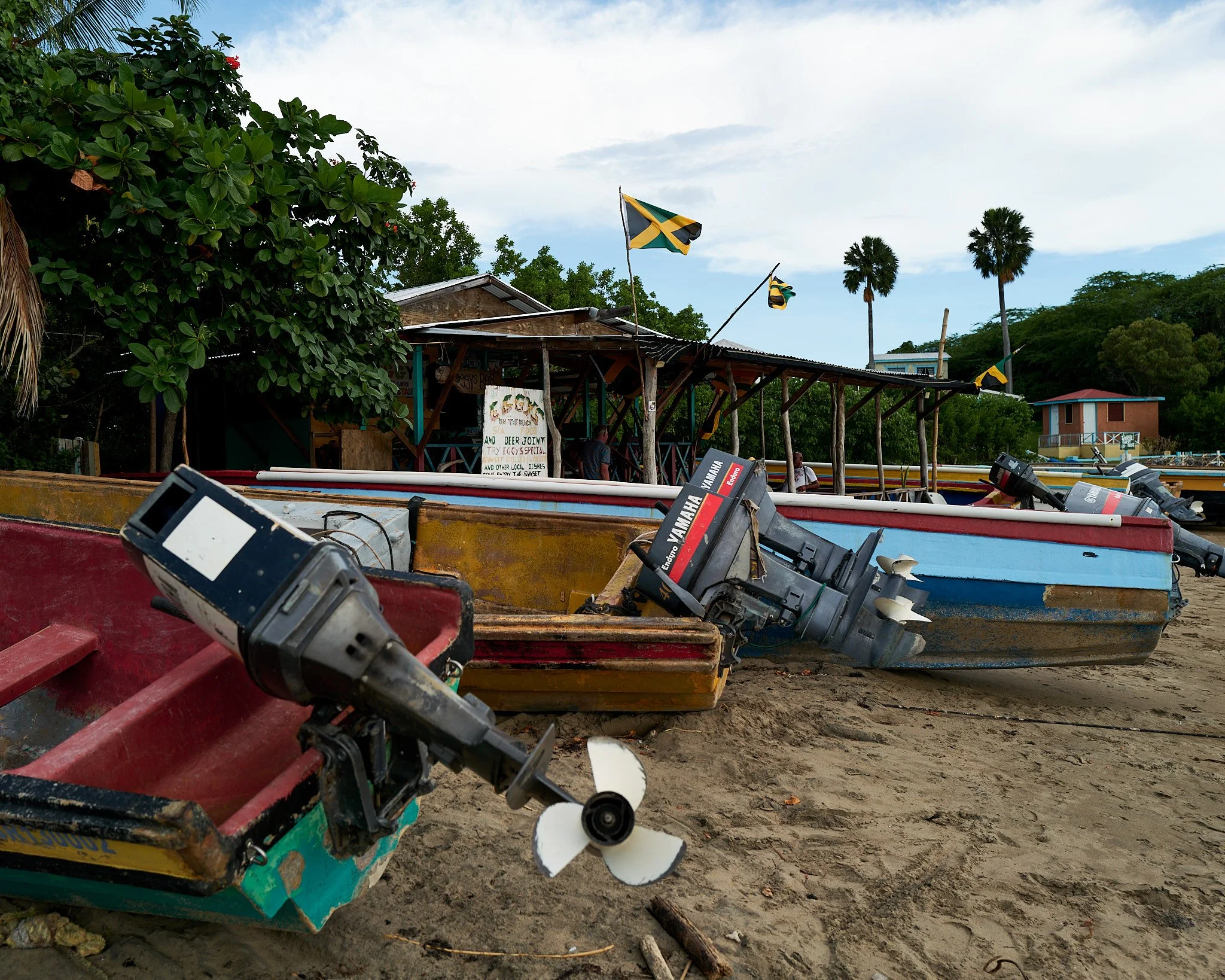 Several small boats with outboard motors on a sandy shore, with flags, a bar or restaurant in the background, and palm trees under a partly cloudy sky.