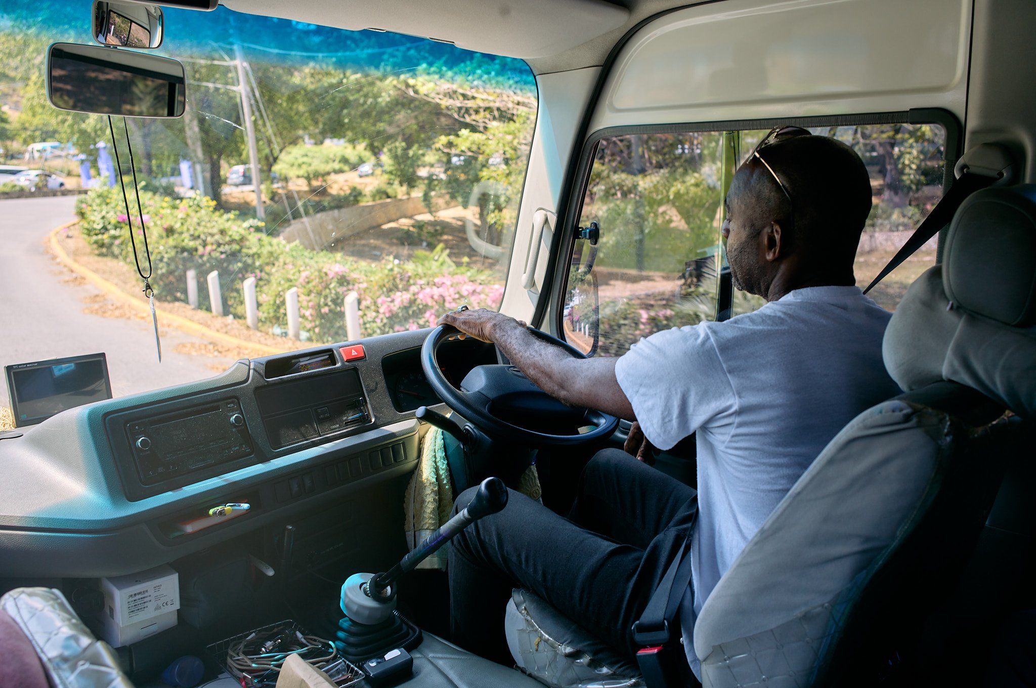 Man driving a vehicle, with a view of the dashboard and road ahead, including greenery and parked cars outside Montego Bay Jamaica