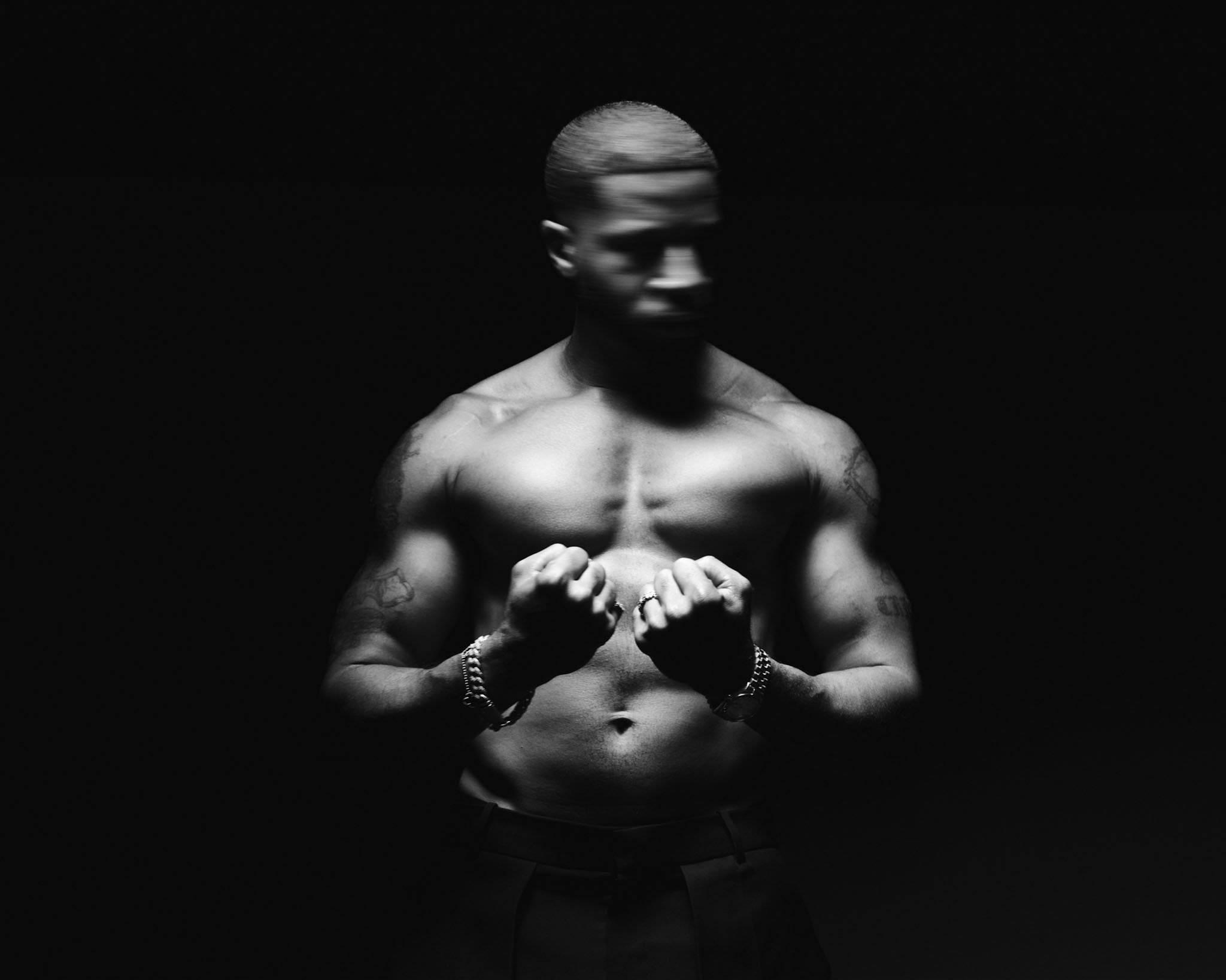 A shirtless man with tattoos, wearing a wristwatch and bracelets, clenches his fists and looks down, with his face blurred and hair styled in a slicked-back manner, against a black background.