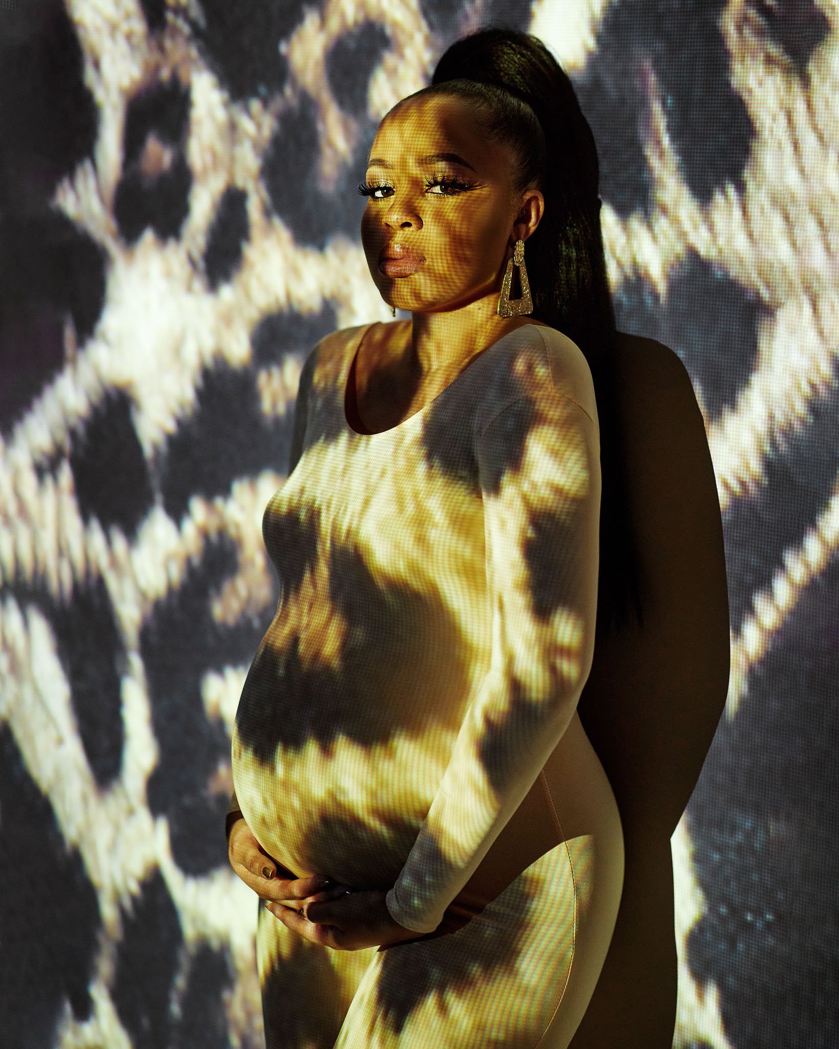 A pregnant woman standing against a patterned background with projection light creating a skull pattern on her and her dress.