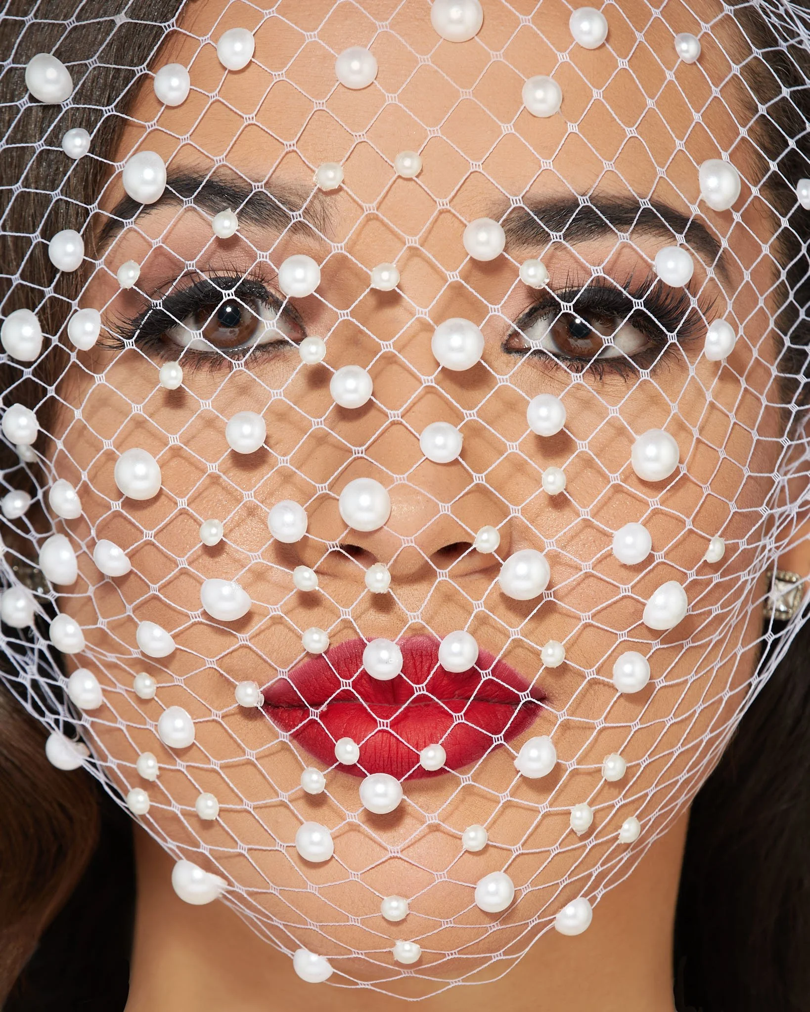 Close-up of a woman's face with makeup, wearing a white pearl-covered netted veil over her face.