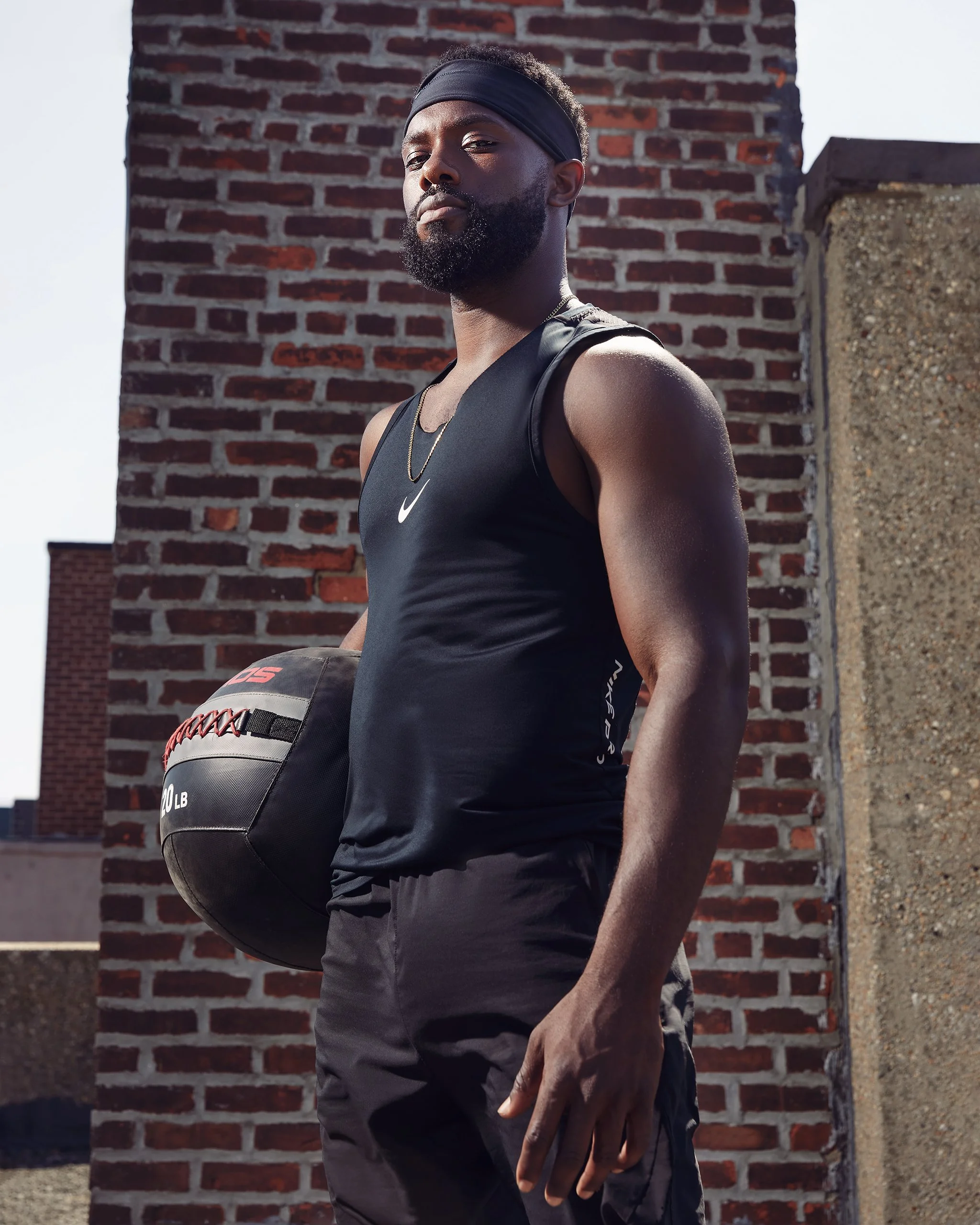 A muscular man with a beard, wearing a black headband, black Nike tank top, and athletic shorts, holding a medicine ball on an outdoor rooftop with a brick wall background.