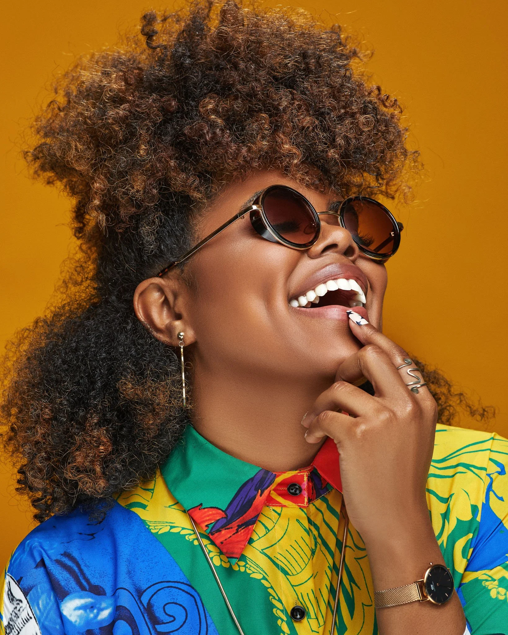 A woman wearing sunglasses, a colorful patterned shirt, and a gold watch, smiling with her hand near her mouth against an orange background.