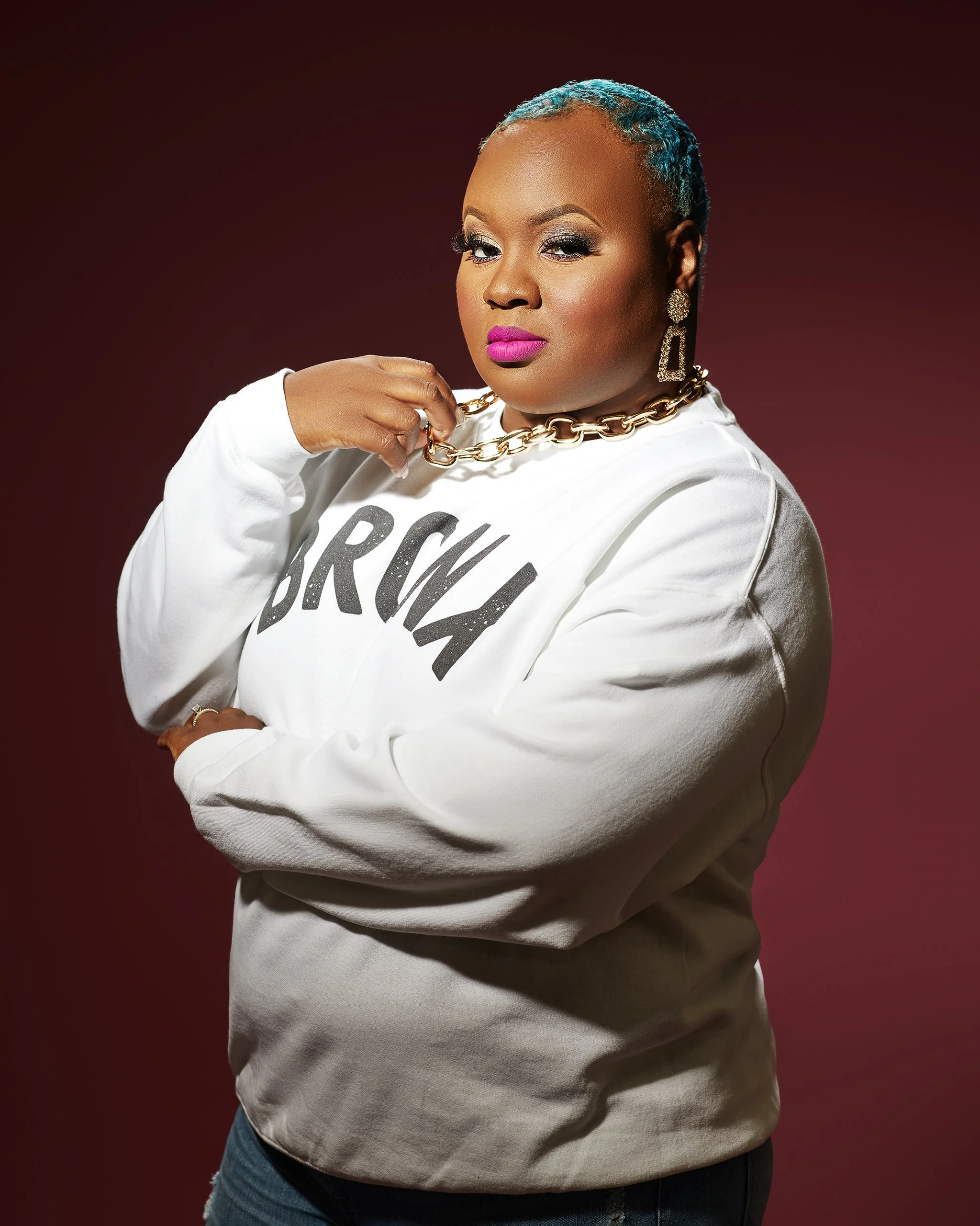 Portrait of a woman with short, dyed blue hair, wearing a white sweatshirt with black lettering, gold jewelry, and bright pink lipstick, posing against a dark red background.