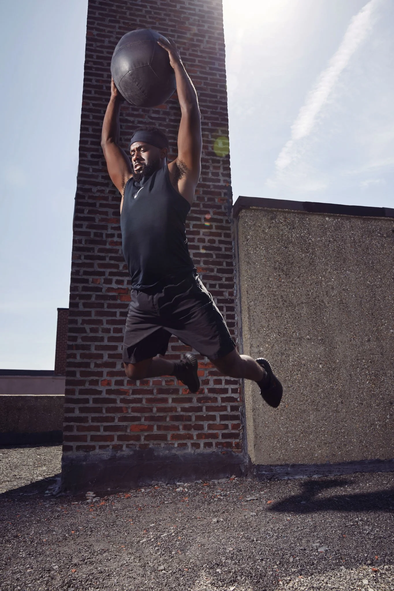 A man wearing black athletic clothing, lifting a medicine ball above his head while jumping on a rooftop with a brick chimney, against a bright sky.