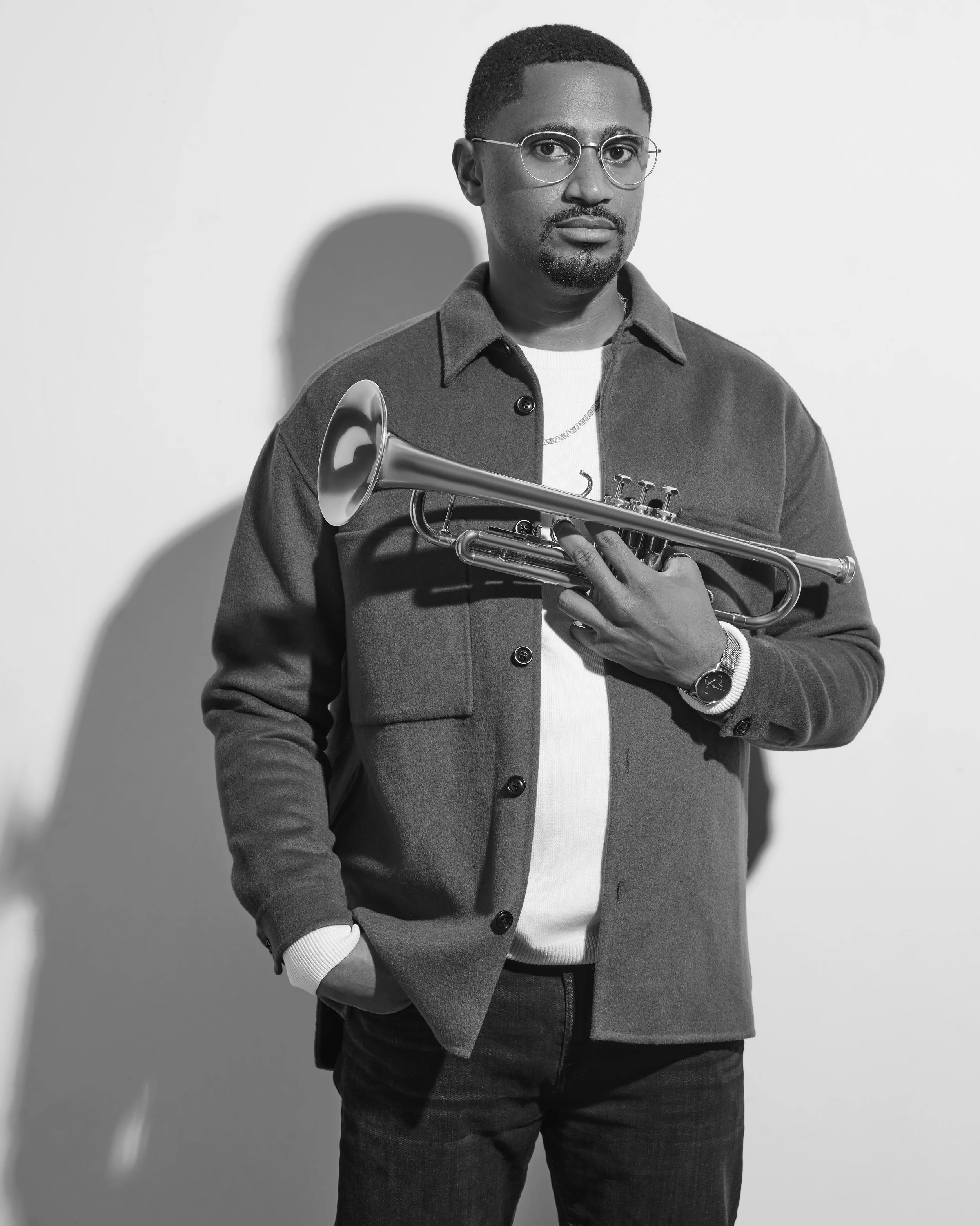 Black and white photo of a man wearing glasses, a white shirt, a watch, and a jacket, holding a trumpet against a plain wall.