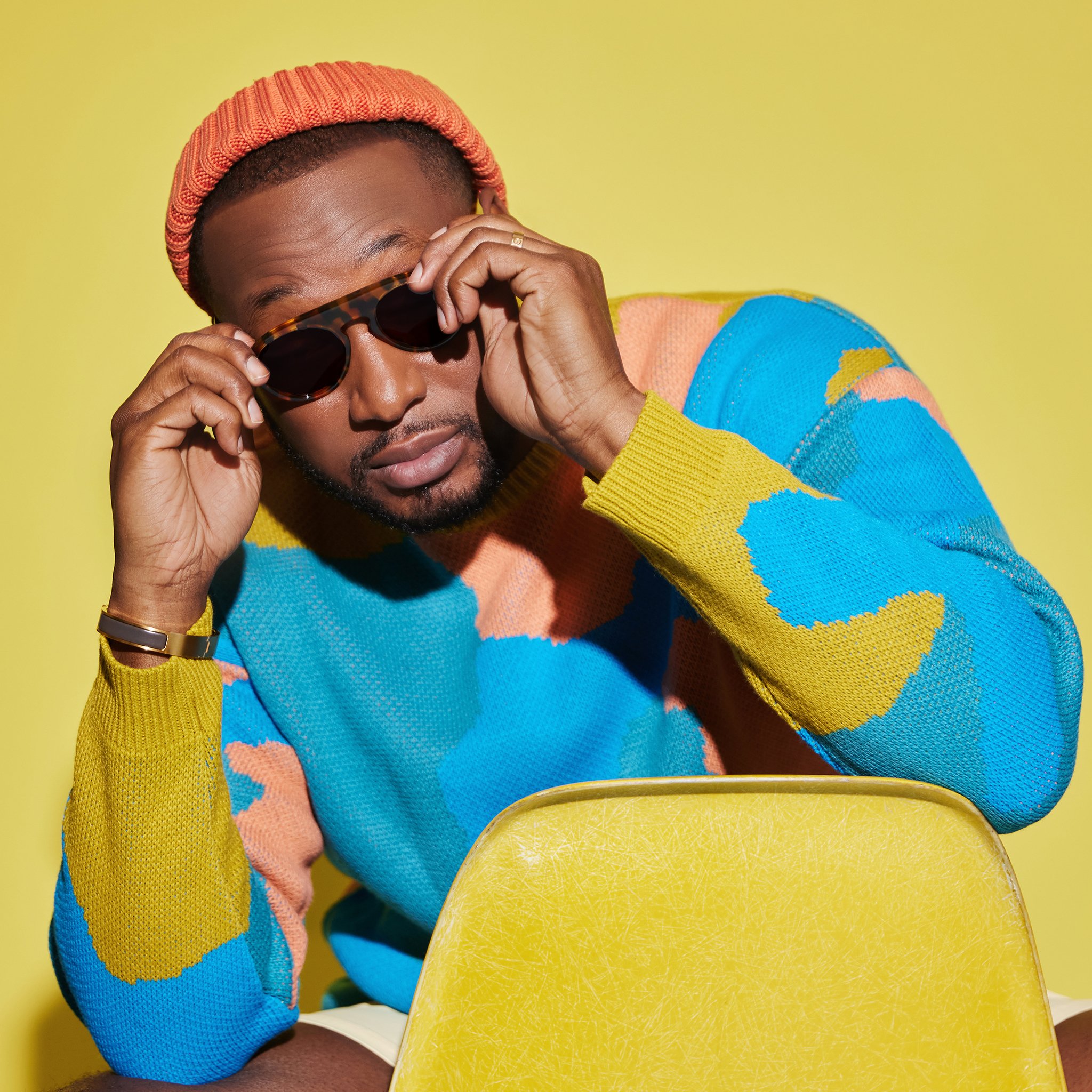 Young man wearing a multicolored sweater, sunglasses, and an orange beanie, sitting at a yellow table against a yellow background.