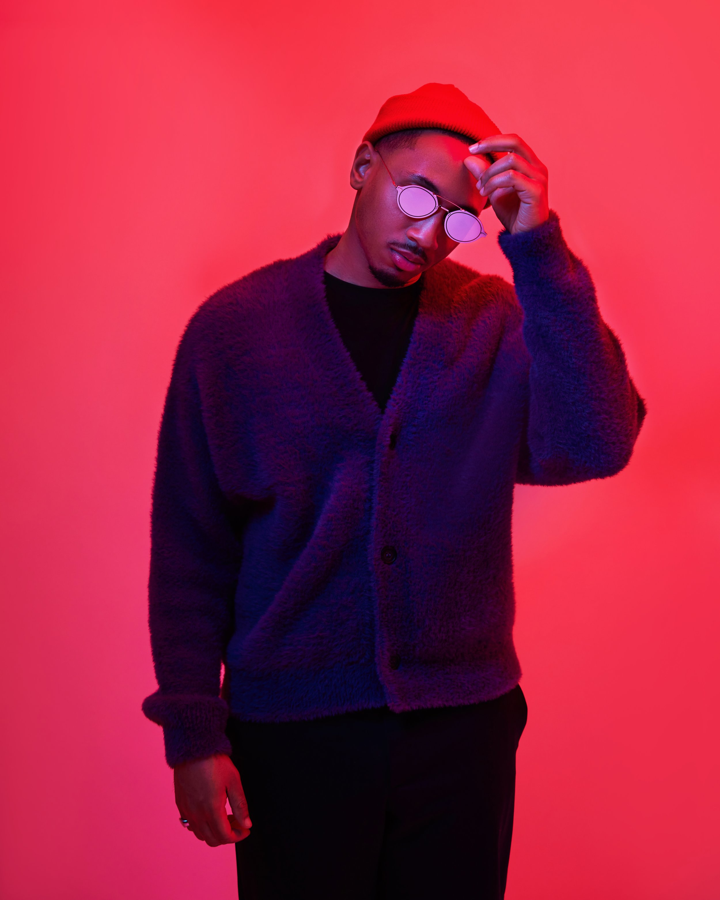 A man wearing a red beanie, sunglasses with round lenses, a black shirt, and a purple fuzzy cardigan posing against a red-pink gradient background.