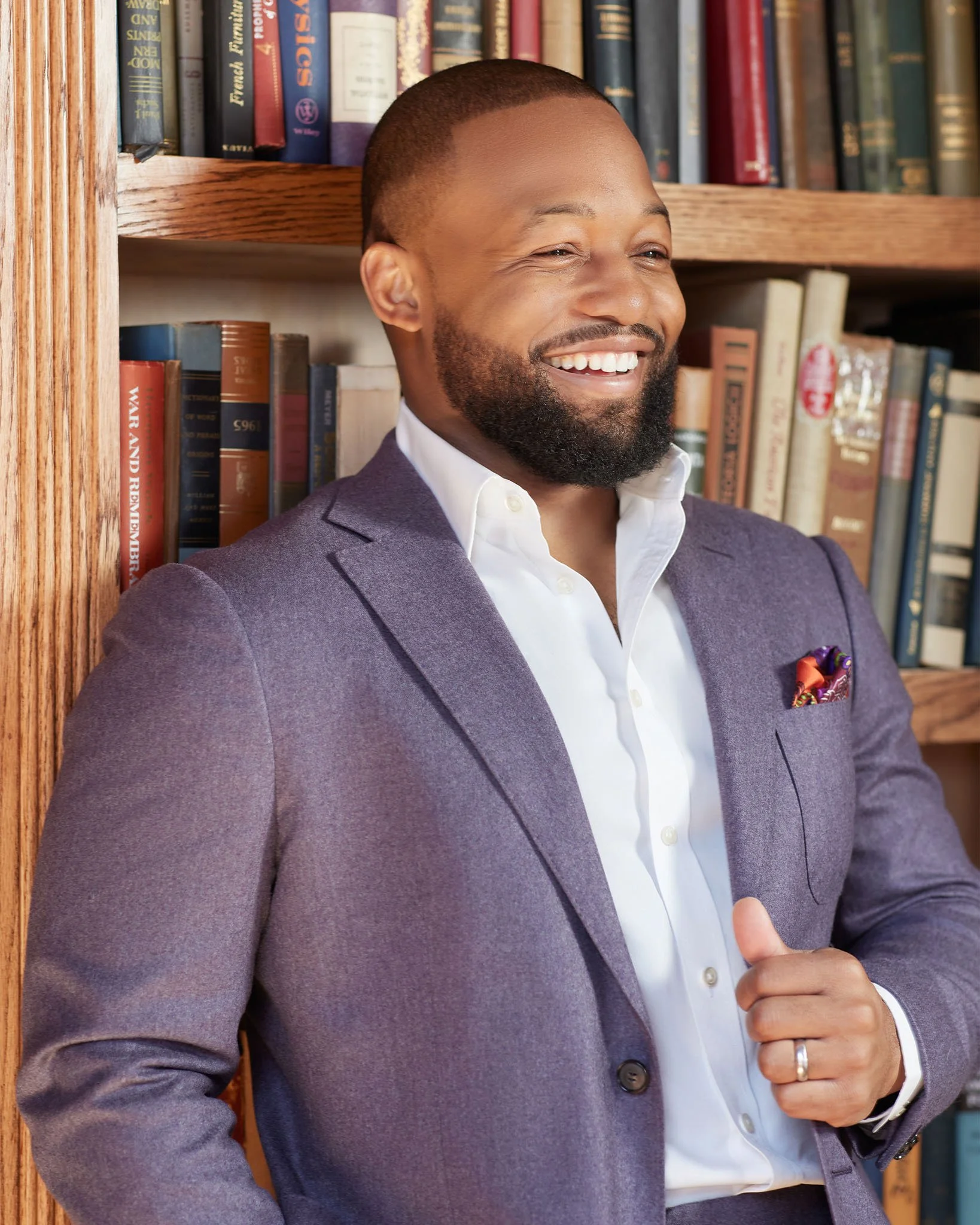 A man with a beard and short hair, smiling, in a purple blazer and white shirt, standing in front of a bookshelf filled with books.