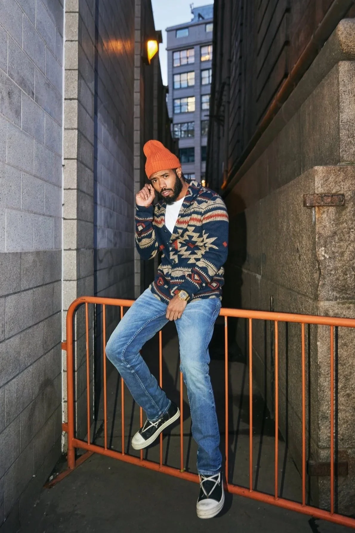A man with a beard and dark hair, wearing an orange beanie, a patterned navy and red sweater, blue jeans, and black and white sneakers, posing in an alleyway at dusk or evening, leaning on an orange barricade with city buildings in the background.