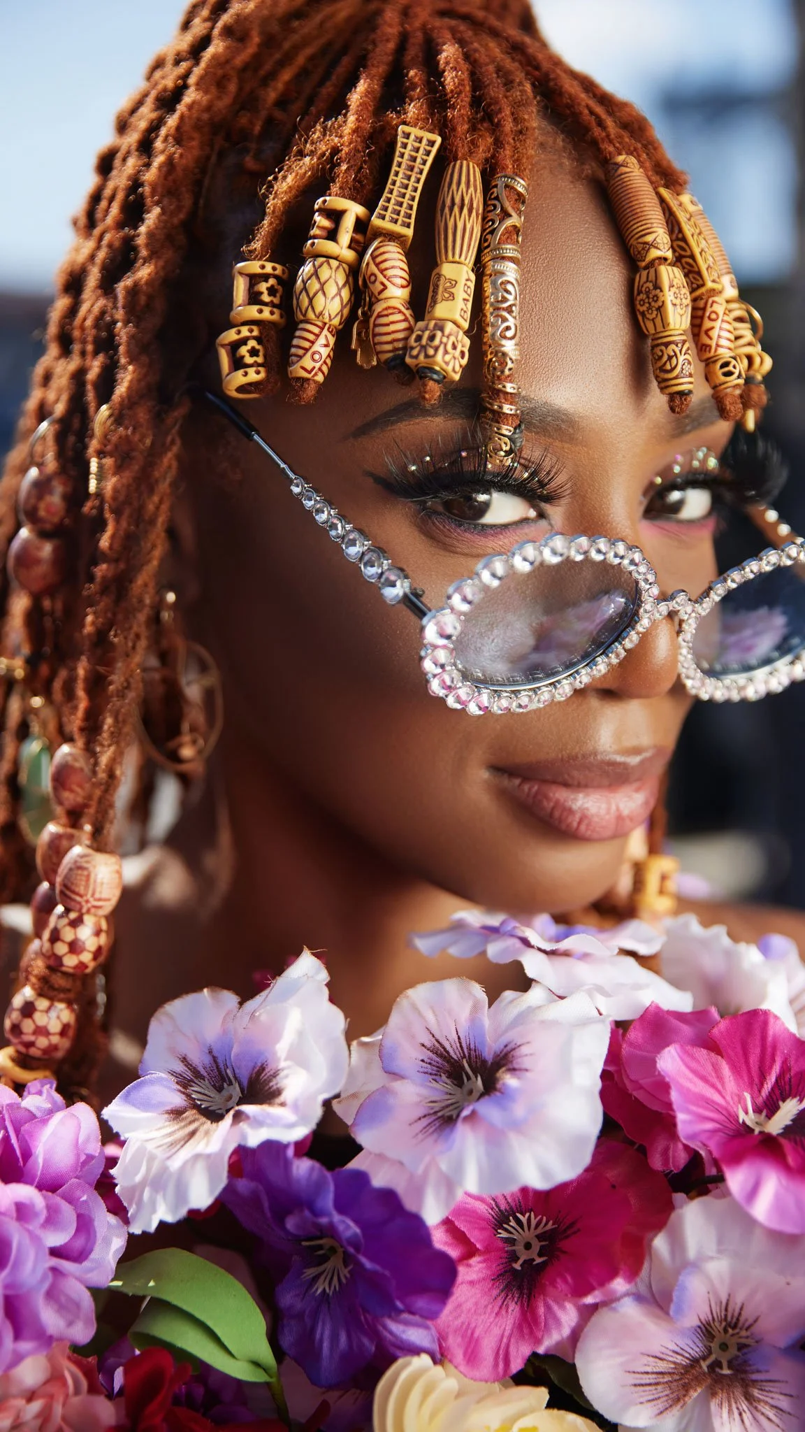 A woman with orange dreadlocks adorned with wooden beads wears large, rhinestone-encrusted sunglasses and is surrounded by colorful flowers.