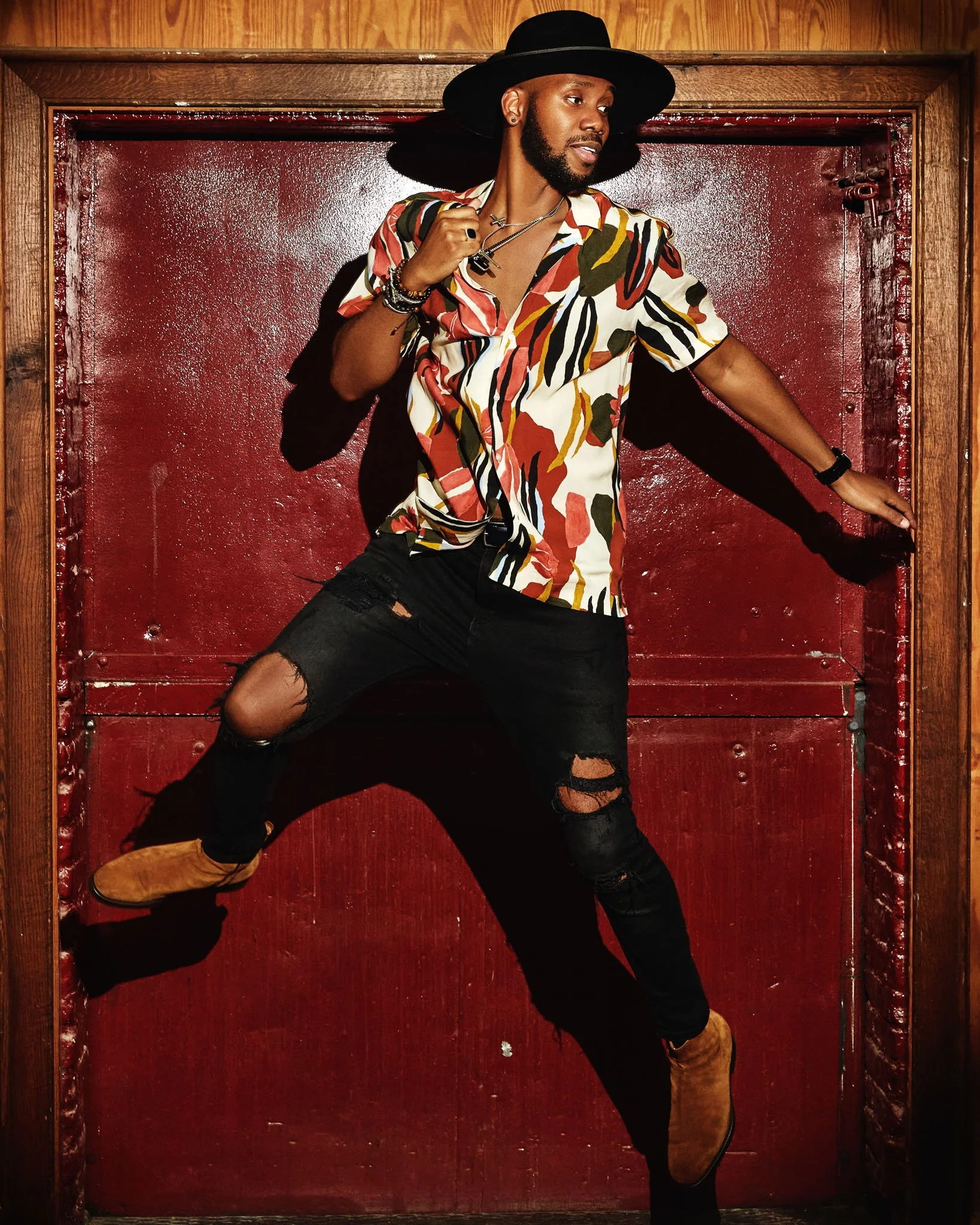 A man with a beard and two necklaces wearing a colorful, patterned shirt, ripped black jeans, tan boots, and a large black hat, is posing against a red textured wall inside a wooden frame.
