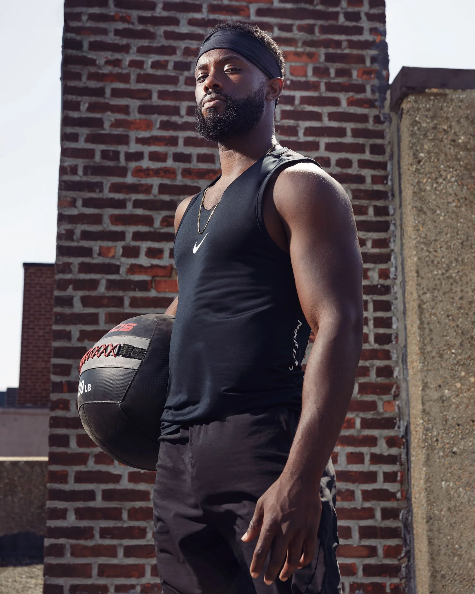 An athletic man standing outdoors next to a brick wall, wearing a black sleeveless workout shirt, black workout pants, a black headband, and a chain necklace. He is holding a medicine ball with red strings and the text '20 LB' on it.