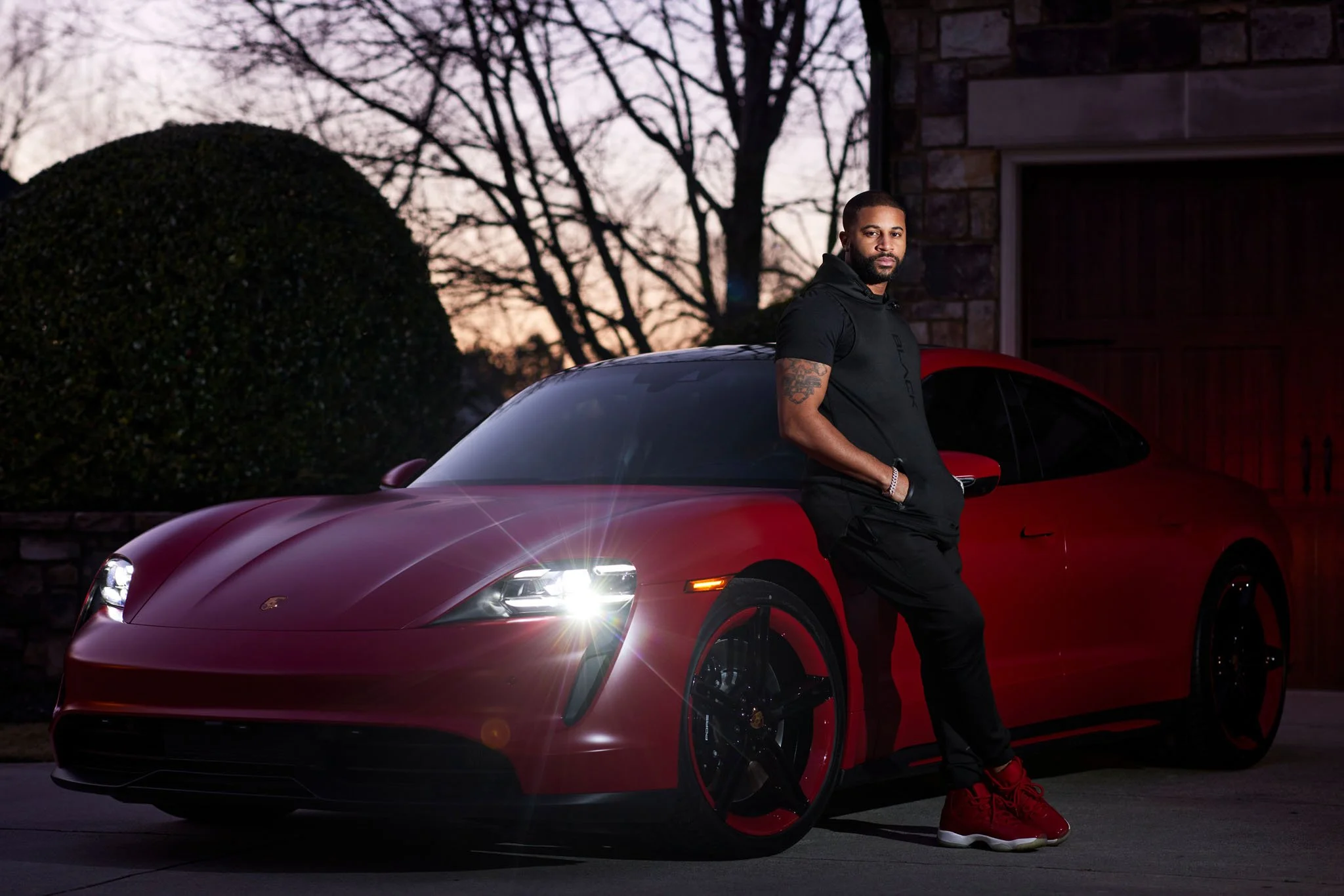 Man with tattoos wearing a black hoodie and black pants standing next to a red sports car with glowing headlights, parked outside during dusk.