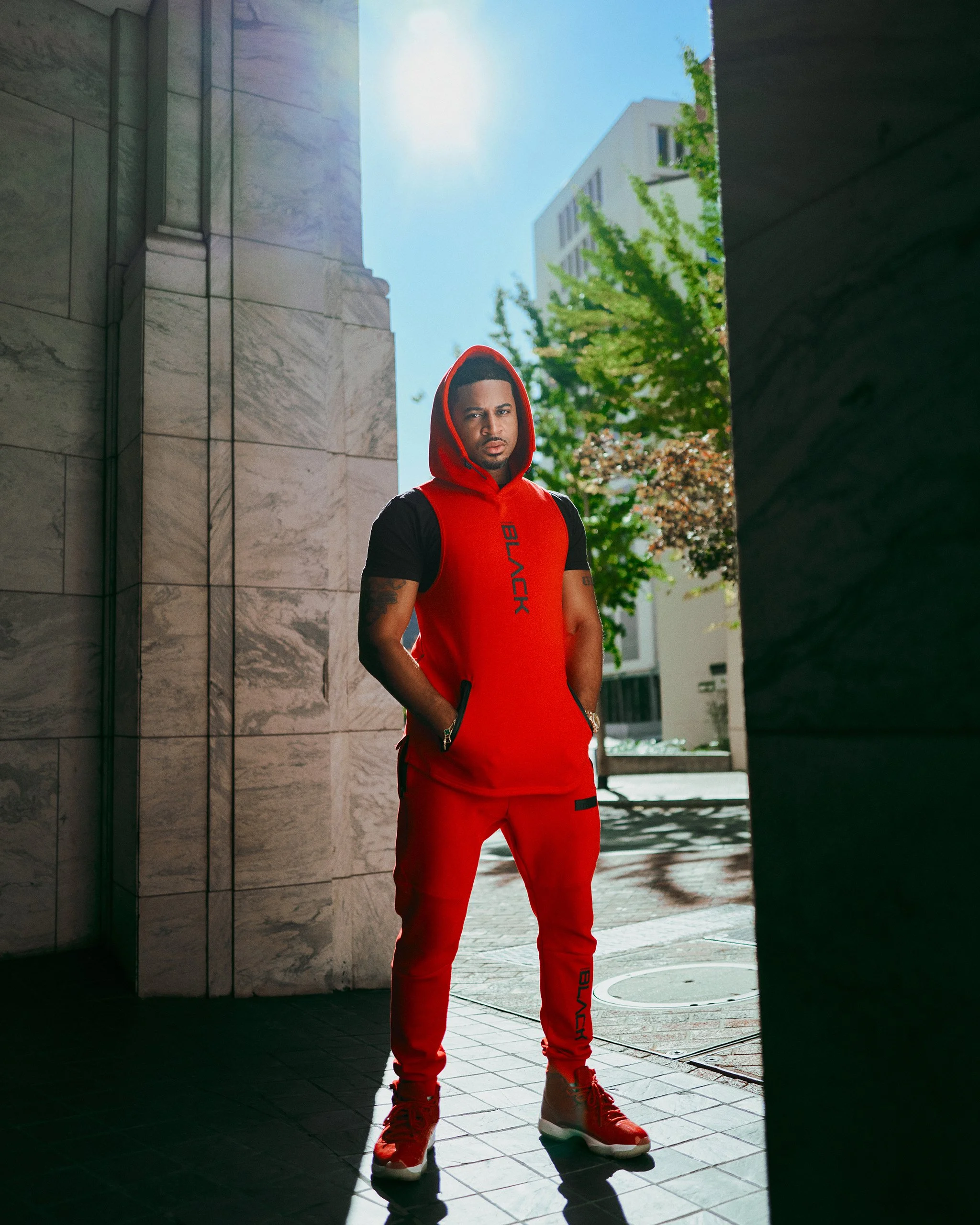 A portrait of Devale Ellis  standing outdoors in a red Actively Black sweatsuit in front of a stone building and trees, during sunny weather.