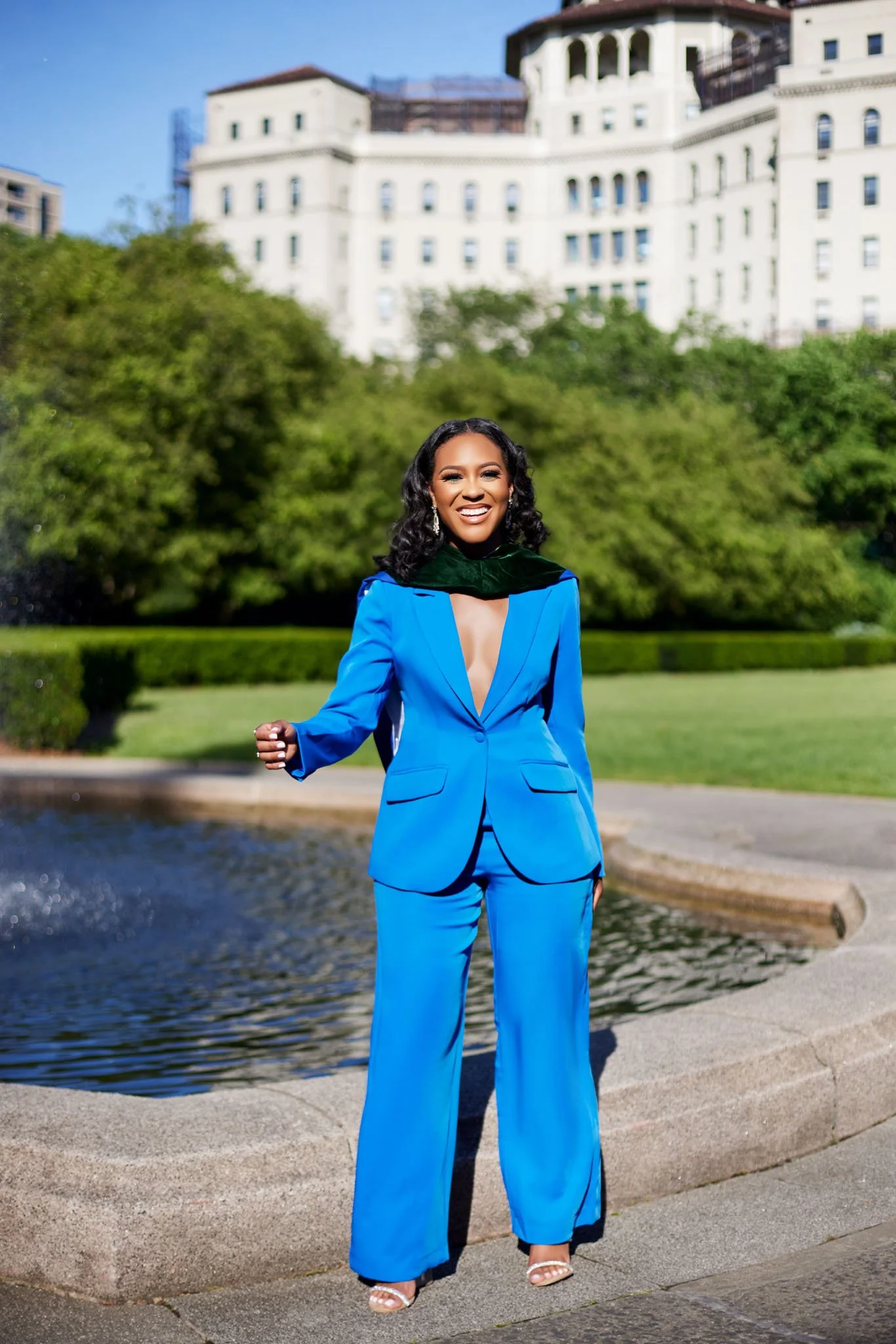 A woman in a bright blue pantsuit with a deep neckline, black scarf, and heels standing outdoors near a fountain, smiling with a background of trees and a large white building.