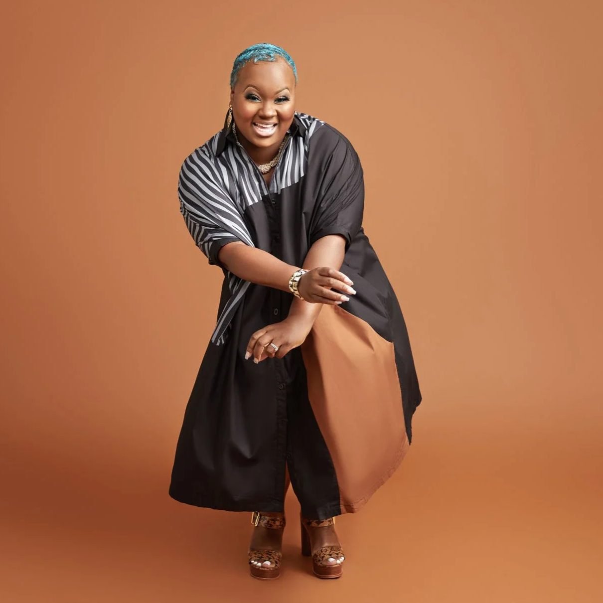 A smiling woman with short blue hair, wearing a black and white striped shirt over a black dress, brown shoes, and gold jewelry, standing against a plain brown background.