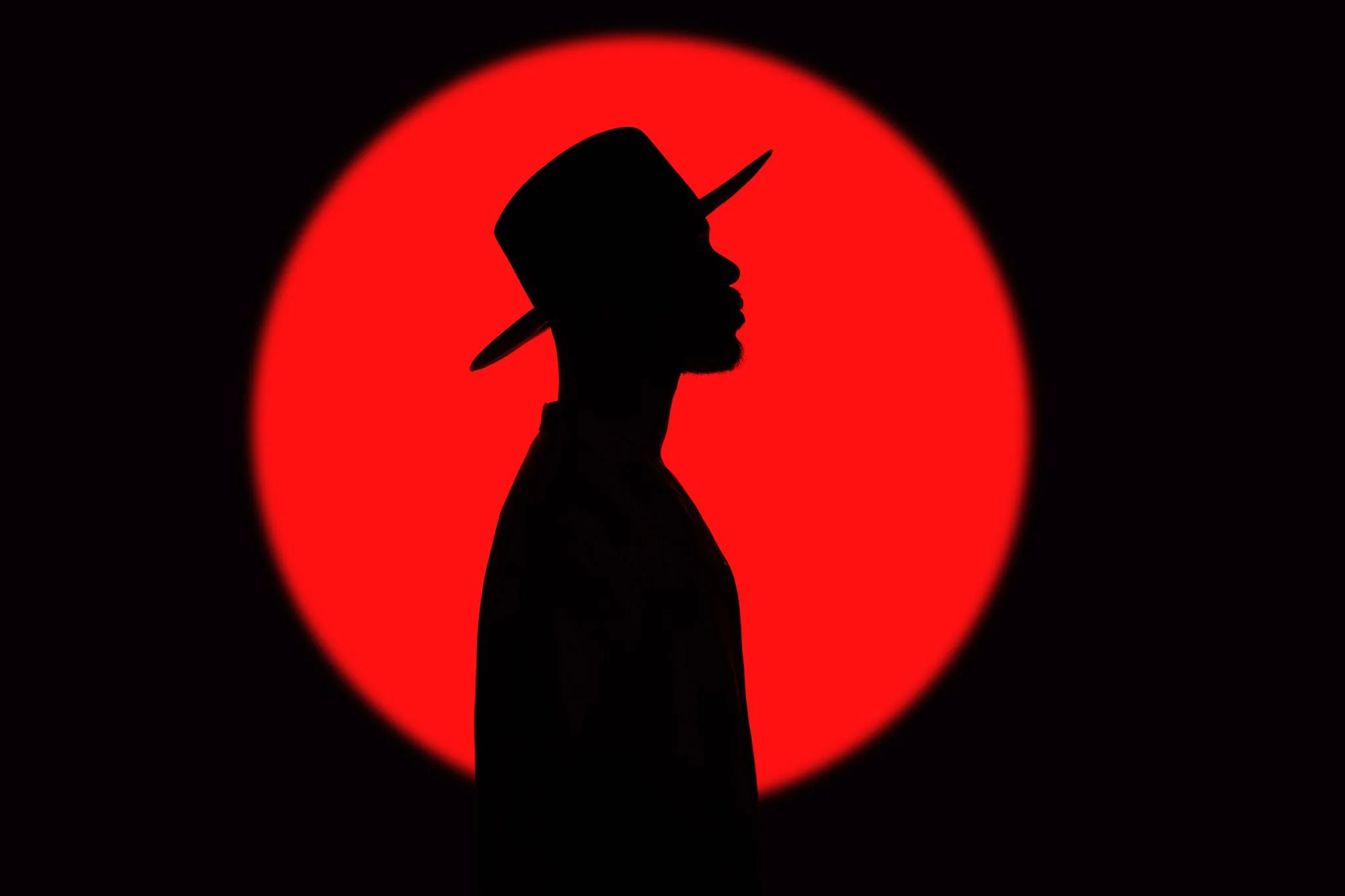 Silhouette of a man wearing a hat with a wide brim, standing in front of a large red circular background, creating a dramatic contrast.