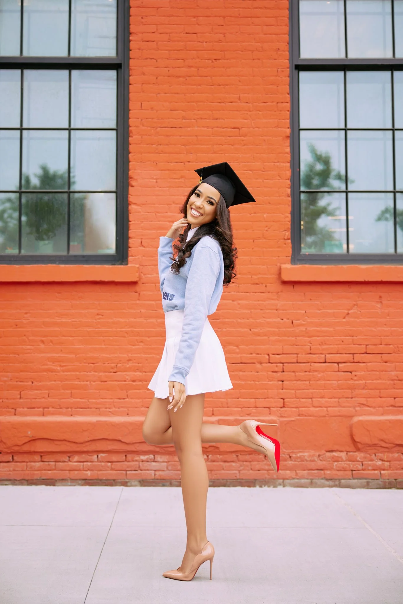 A young woman in a graduation cap and casual outfit standing in front of a red brick building, smiling and balancing on one leg with the other leg bent.