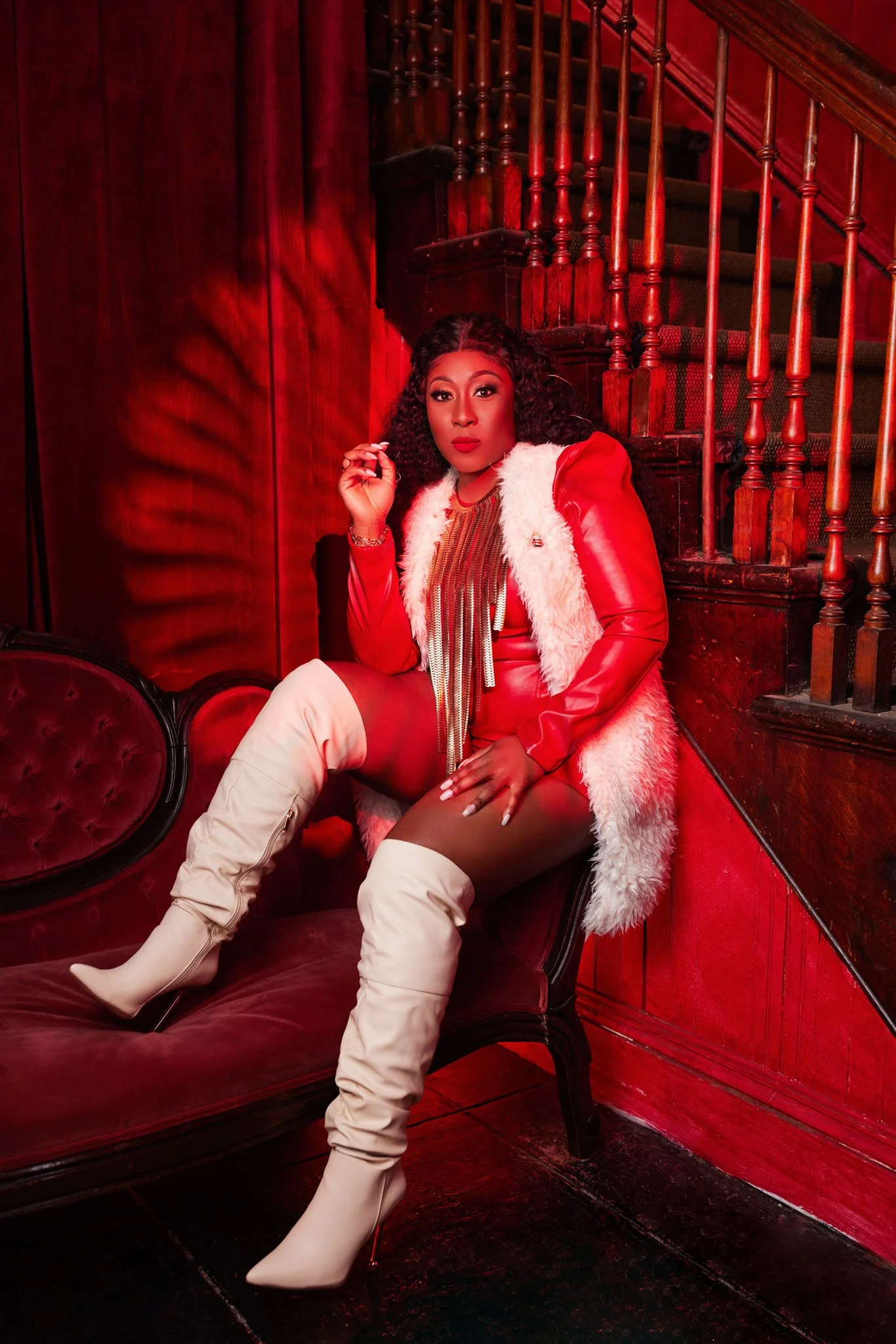 Woman seated on vintage red velvet sofa, wearing a red jacket with white fur trim, a striped top, and thigh-high beige boots in a dimly lit, wood-paneled room with a staircase in the background.