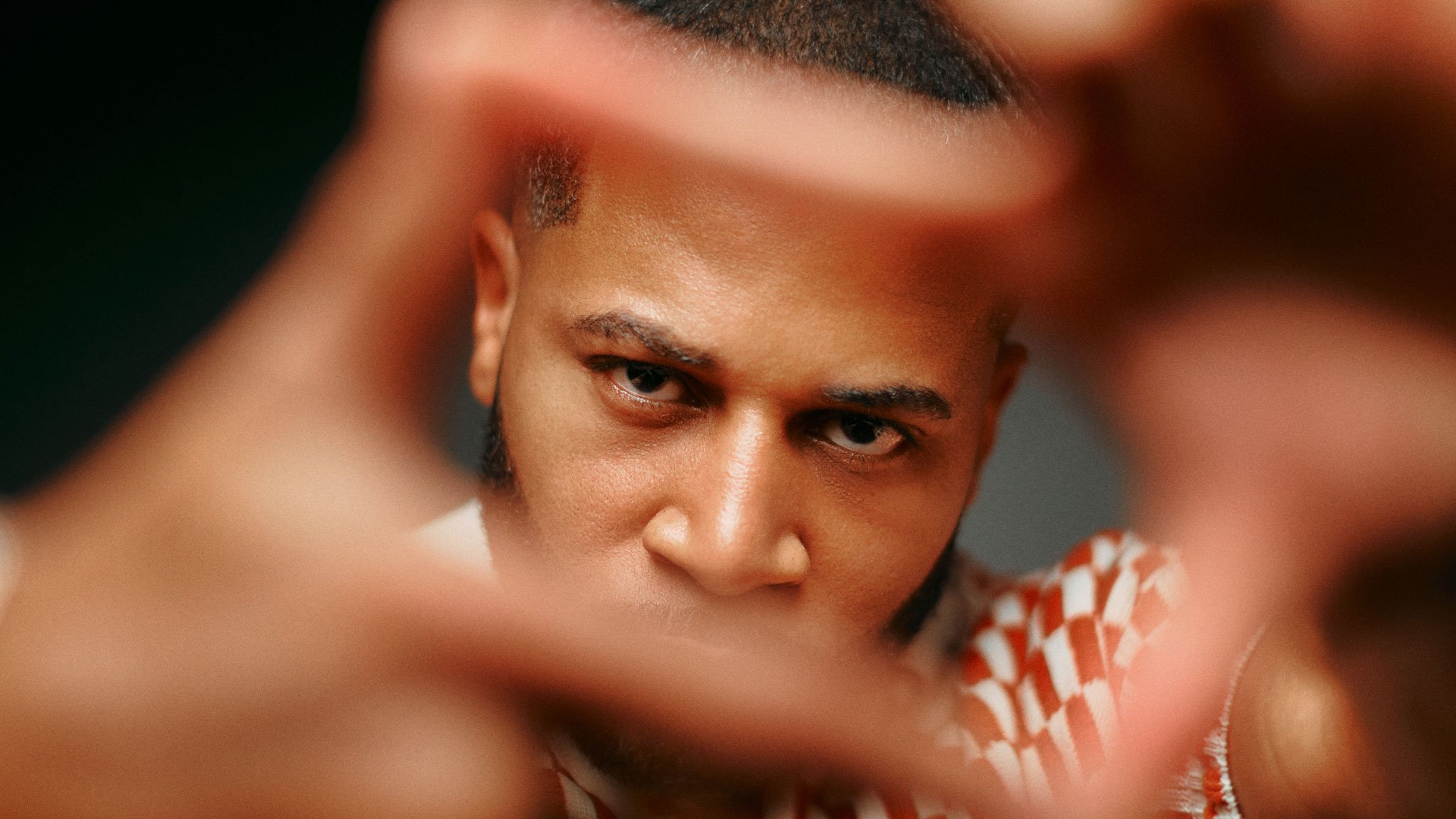 Close-up portrait of a young man looking intently through a frame made by his hands, with a confident expression and direct gaze.