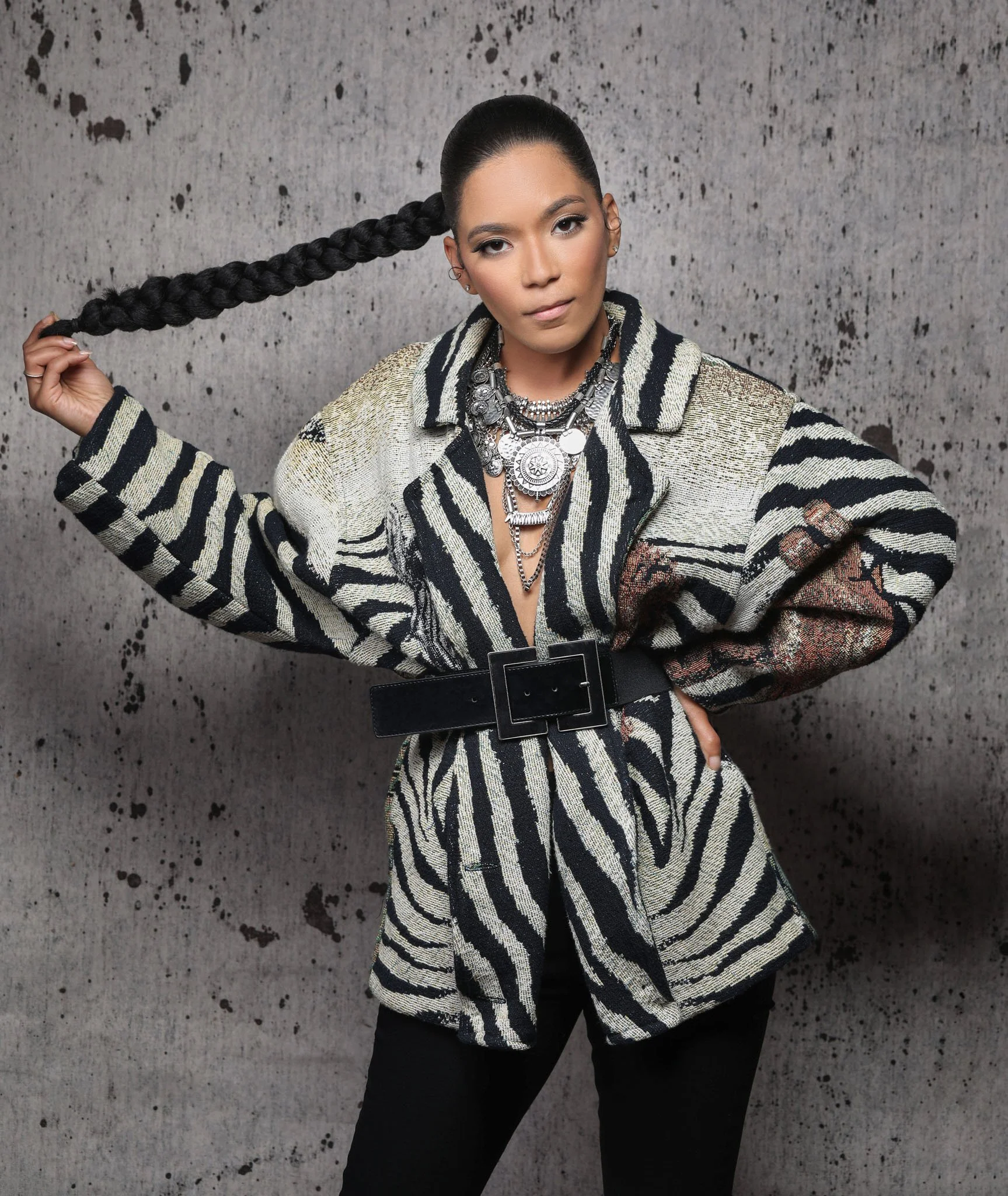 Fashion model wearing a black, white, and beige zebra print outfit with multiple necklaces, standing in front of a textured gray wall.