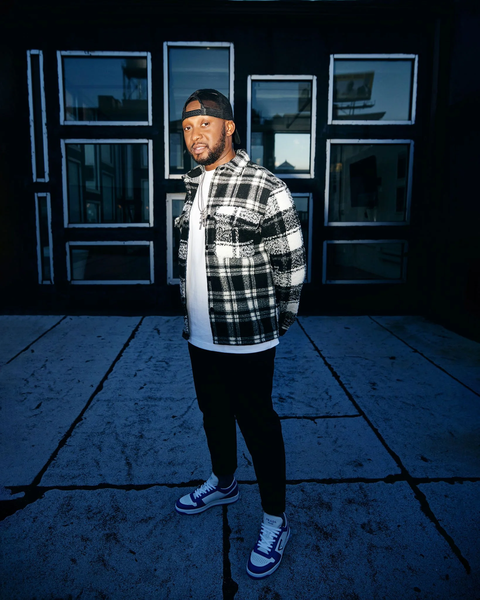 A man standing on a concrete floor outdoors in front of a black wall with geometric window designs, wearing a black and white plaid jacket, white t-shirt, black pants, blue and white sneakers, and a black cap worn backwards.