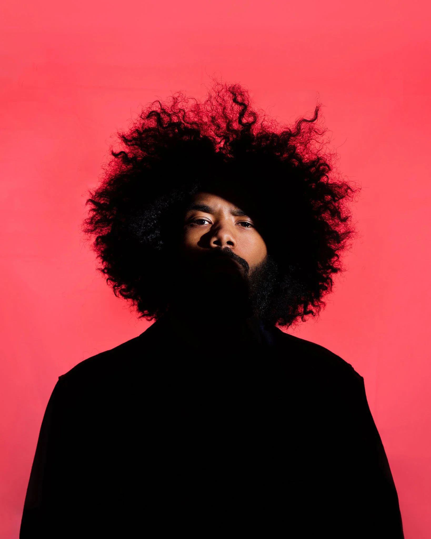 A man with large curly black hair and a beard, wearing a black shirt, posing against a pink background.