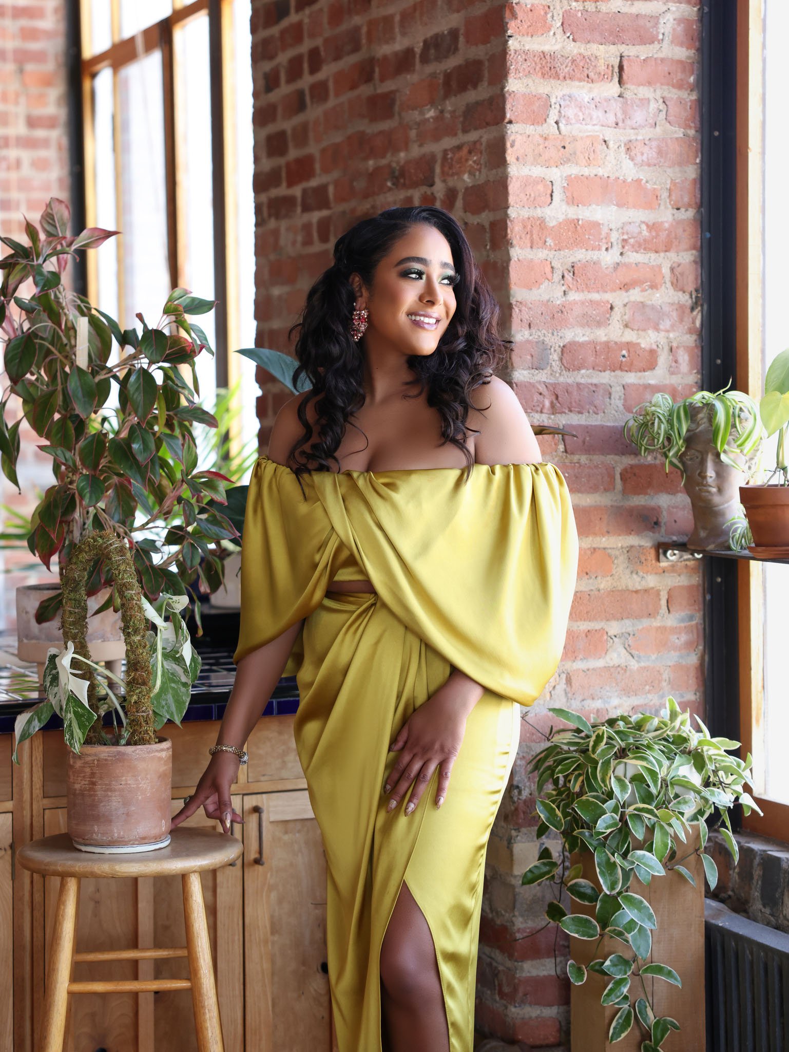 A woman with dark curly hair, wearing a yellow off-the-shoulder gown with a thigh-high slit, stands near a brick wall and large windows, surrounded by houseplants, smiling and looking to her right.