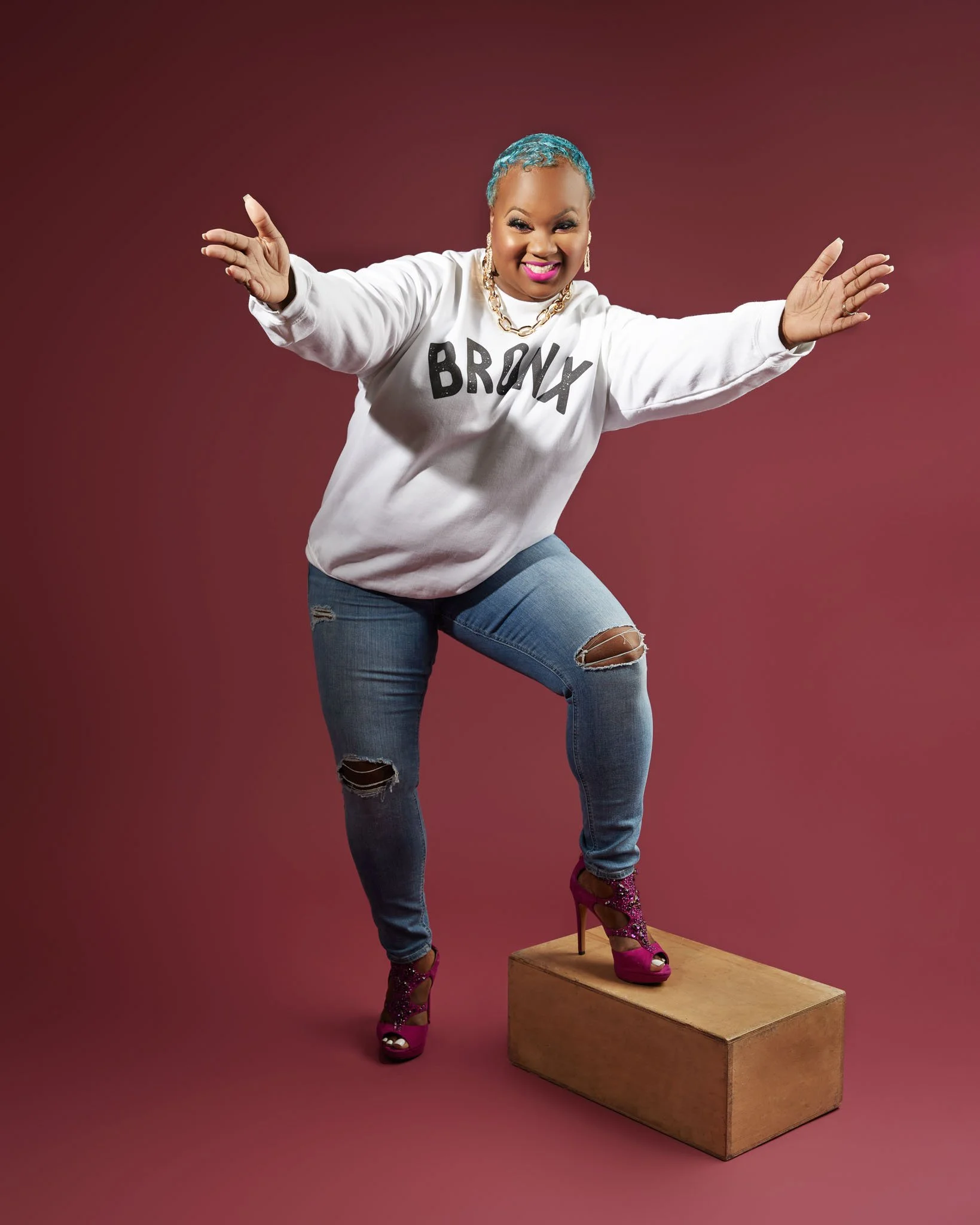 Woman with short blue hair posing confidently with one foot on a wooden block, wearing a white sweatshirt with 'BRONX' written on it, ripped jeans, and pink high heels, against a pink background.
