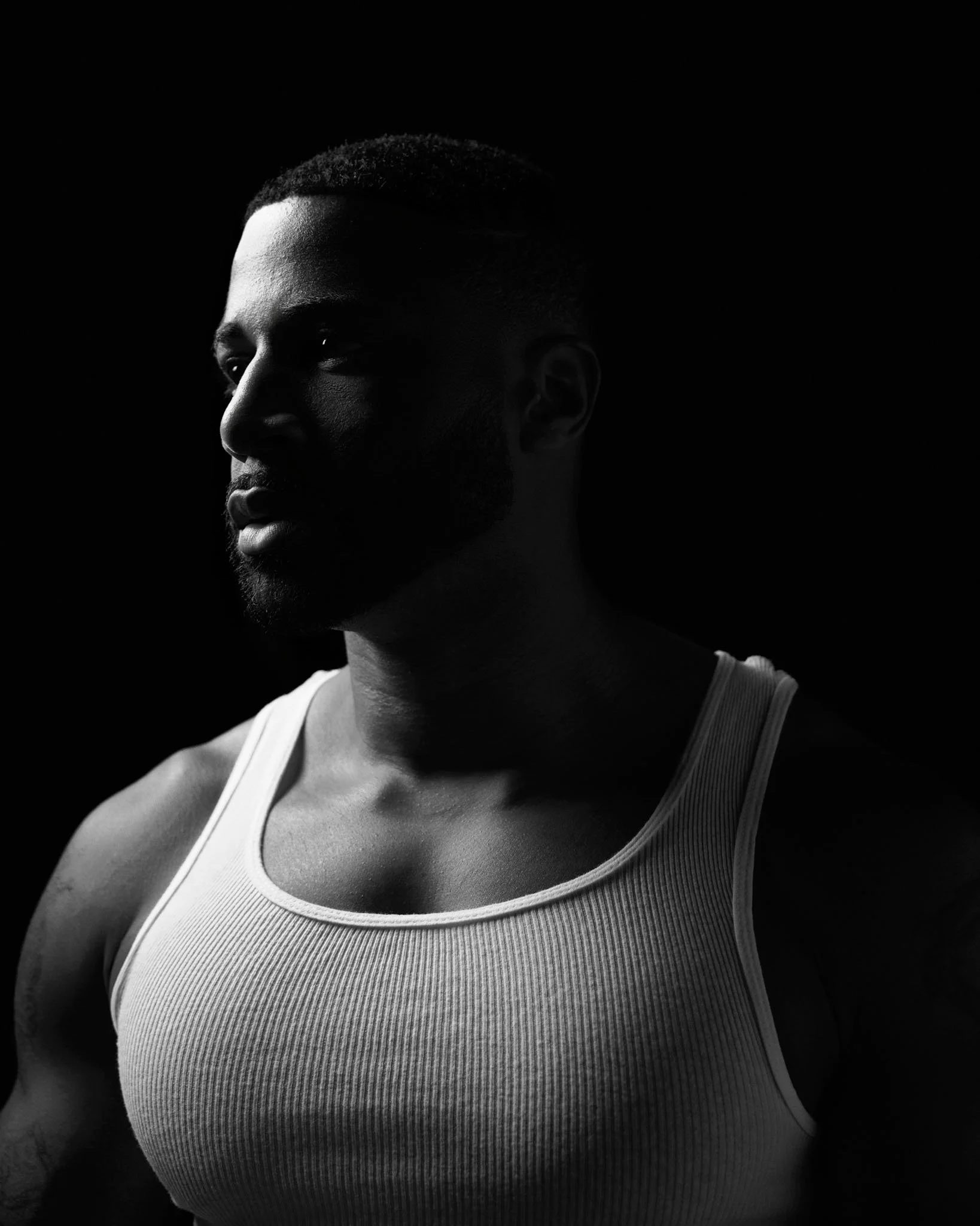 A black and white portrait of a muscular man wearing a sleeveless shirt, looking to the side with a serious expression, against a dark background.